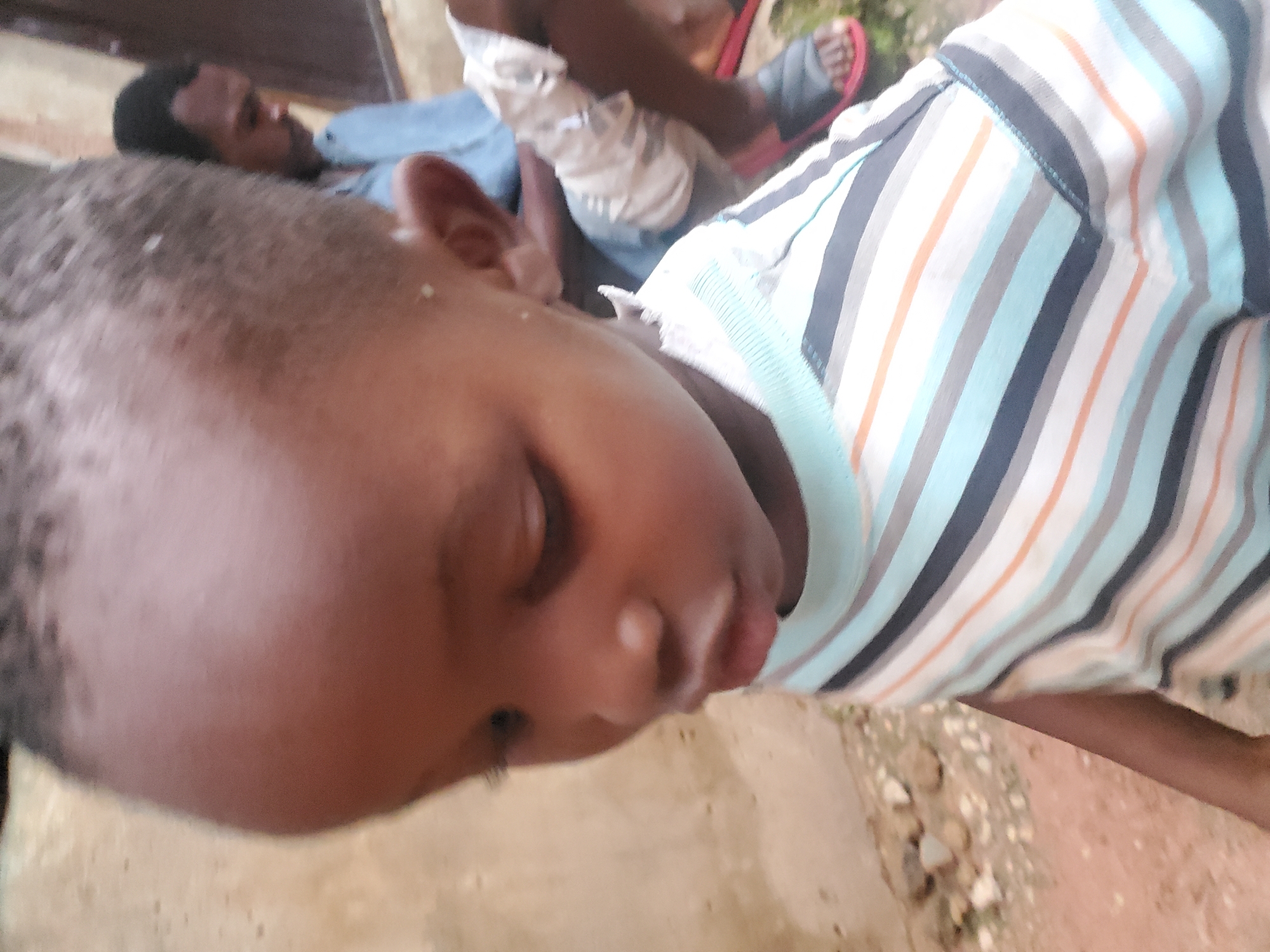 The image depicts a close-up, slightly blurred view of a young Black child in the foreground, facing downwards and to the left. The child is wearing a striped t-shirt with horizontal bands of light blue, white, grey, and orange. Their head is tilted, and their eyes are not clearly visible. The background features a blurry, light-colored wall and some ground with small stones. To the right of the child, in the background, a second, adult Black male is visible, also out of focus. This adult is wearing a light blue shirt. The environment appears to be outdoors, possibly in a residential area. The overall lighting suggests daylight. This image was captured in Jos, Nigeria.