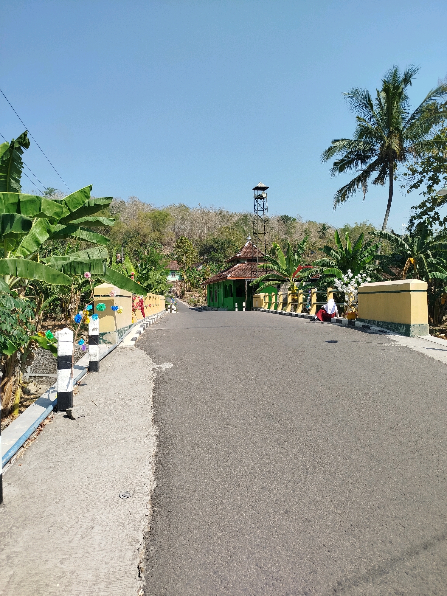 The image depicts a rural road in Semin, Indonesia, on a clear, sunny day. The road curves to the right, leading towards a small mosque with a green roof and walls, and a tall, dark tower next to it. Banana trees with large, vibrant green leaves line the left side of the road, and coconut palm trees are visible in the background. A yellow wall with decorative elements runs along the right side of the road. A person wearing a white hijab and a red skirt is sitting on the ground near the yellow wall, facing the mosque. The road is paved, and a concrete sidewalk runs along the left side, bordered by a low stone wall. The background shows a hilly landscape with dry, sparse vegetation and some scattered houses.