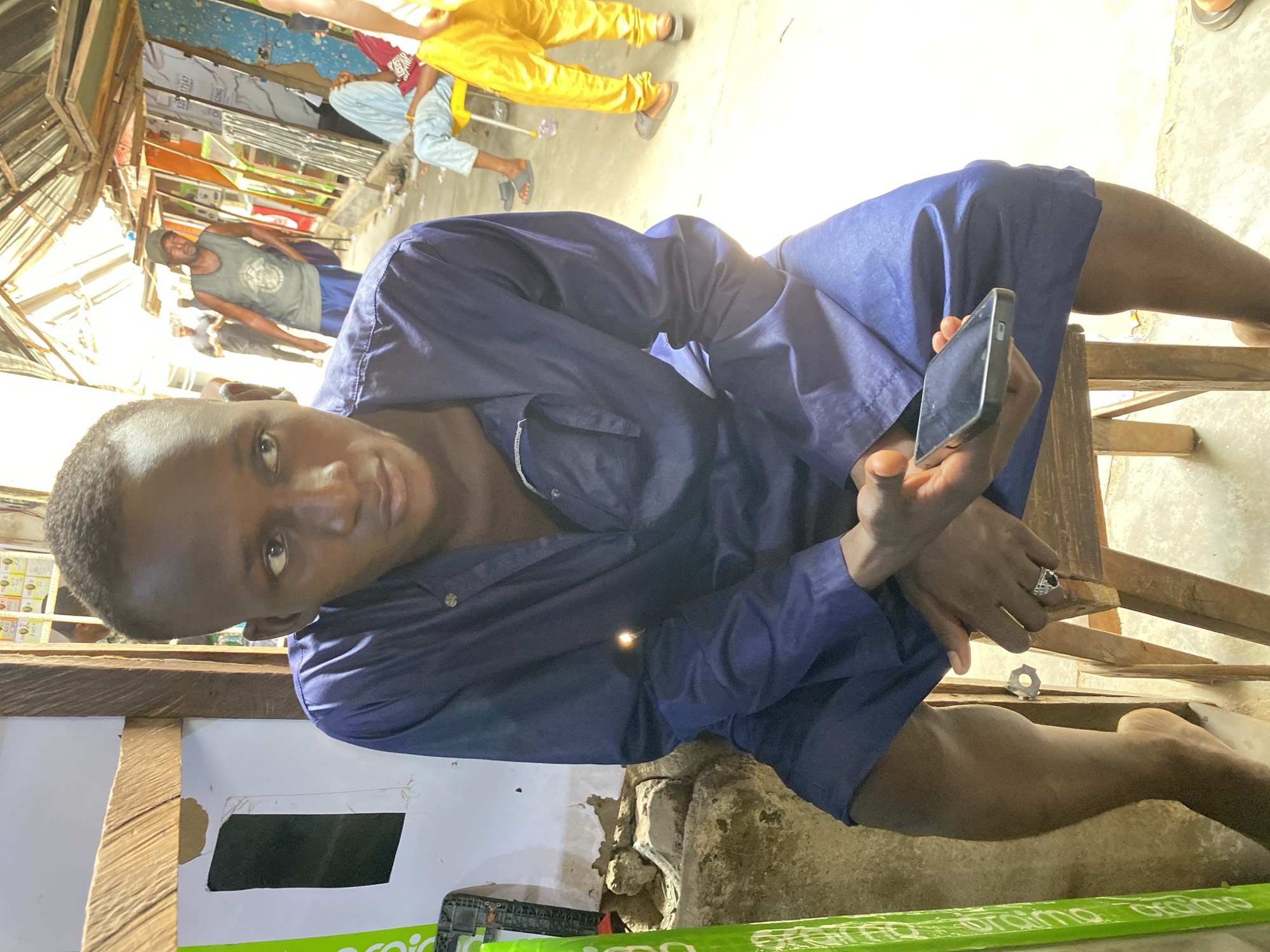 A young man in a dark blue button-down shirt sits on a wooden chair, looking at the camera while holding a smartphone in his hand. The setting appears to be a roadside shop or market in Lokoja, Nigeria. Other individuals are visible in the background, including a man in a grey tank top. The area is cluttered with various objects and displays of goods. The environment shows signs of being an informal, open-air commercial area.