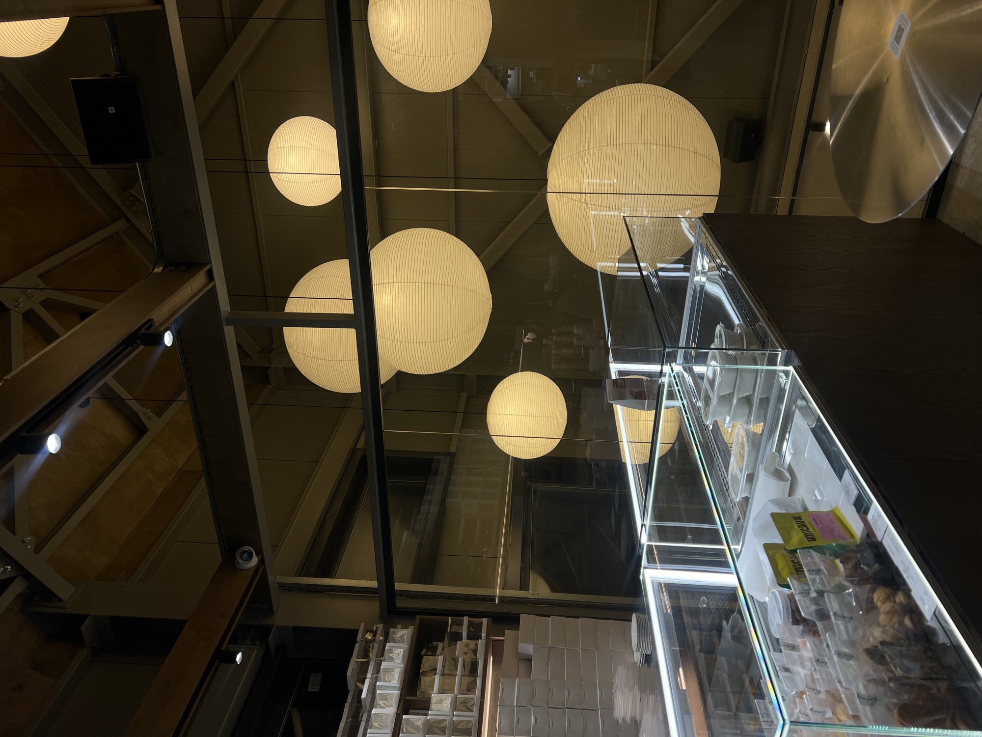 The image depicts an interior setting, likely a commercial space, as indicated by the presence of a glass display case filled with packaged goods. The display case is illuminated by bright LED strips and contains items such as packaged food, possibly cookies, or snacks. Above, the ceiling is fitted with a network of metal beams and ductwork, punctuated by multiple round paper lanterns providing ambient lighting. A black speaker hangs from the ceiling, suggesting the space may be a cafe or shop with an audio system. The items stacked below, along with the reflective surfaces throughout the space, suggest that this is some sort of shop.
While the image doesn't provide visual cues to pinpoint the location in Vladivostok, Russia, if it is in that city, it would likely be in a modernized commercial or shopping area. The image doesn't contain any people or interactions.