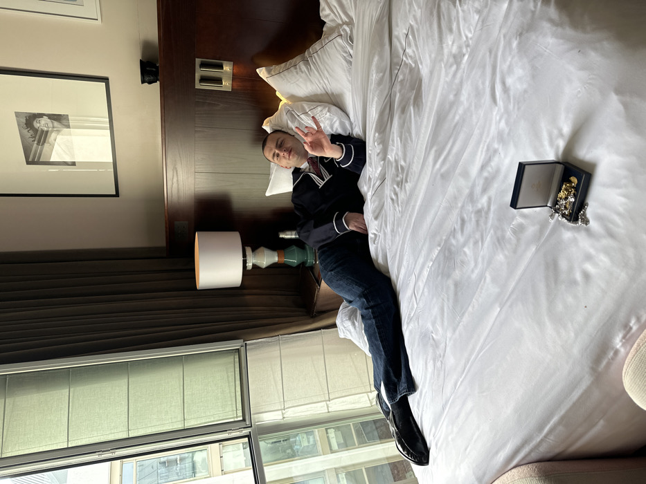 The image displays an interior scene within a hotel room located in Paris, France. A male individual, appearing to be in his 20s or 30s, is positioned lying on his left side on a large bed covered with white linens and a white duvet. He is wearing a dark blue long-sleeved shirt featuring white trim on the collar and cuffs, dark blue jeans, and black flat slip-on shoes. His head rests on a white pillow, and he looks directly at the camera with a neutral expression. His right hand is raised, forming a two-finger "peace" or "victory" gesture.
To the left of the bed, a wooden bedside table supports an illuminated lamp with a white cylindrical shade and a distinct multi-colored base. A large window with partially drawn light-colored fabric shades reveals an exterior view of light-toned urban buildings. Dark, vertically pleated curtains are drawn to the right of the window frame. Above the dark wood headboard, a framed black and white photograph of a woman's face is mounted on the wall. A dark, ornate decorative object rests on the headboard's shelf.
On the white duvet, near the lower right portion of the image, an open dark blue rectangular jewelry box is present. The interior of the box is light-colored, and the brand name "Cartier" is visible on the inside lid. The box contains various items of jewelry, including several gold-colored chains and pendants, with a silver-colored chain or necklace partially extended outside the box. A portion of a light-colored upholstered chair or sofa is visible in the bottom left corner.