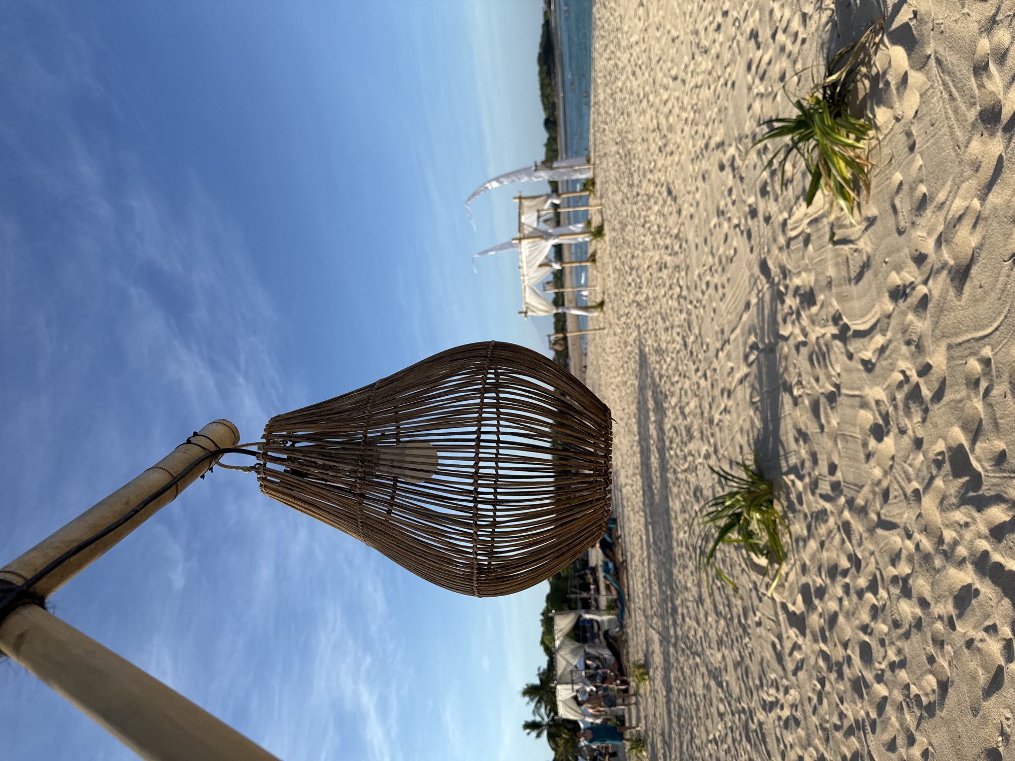 The image shows a beach scene in Kuta Selatan, Indonesia. In the foreground, a teardrop-shaped rattan lampshade hangs from a bamboo pole. The background features white sand, vegetation, and several white canopies with flowing fabric, suggesting a lounge or seating area. Distant figures of people are discernible. The sky above is clear blue with wispy clouds.