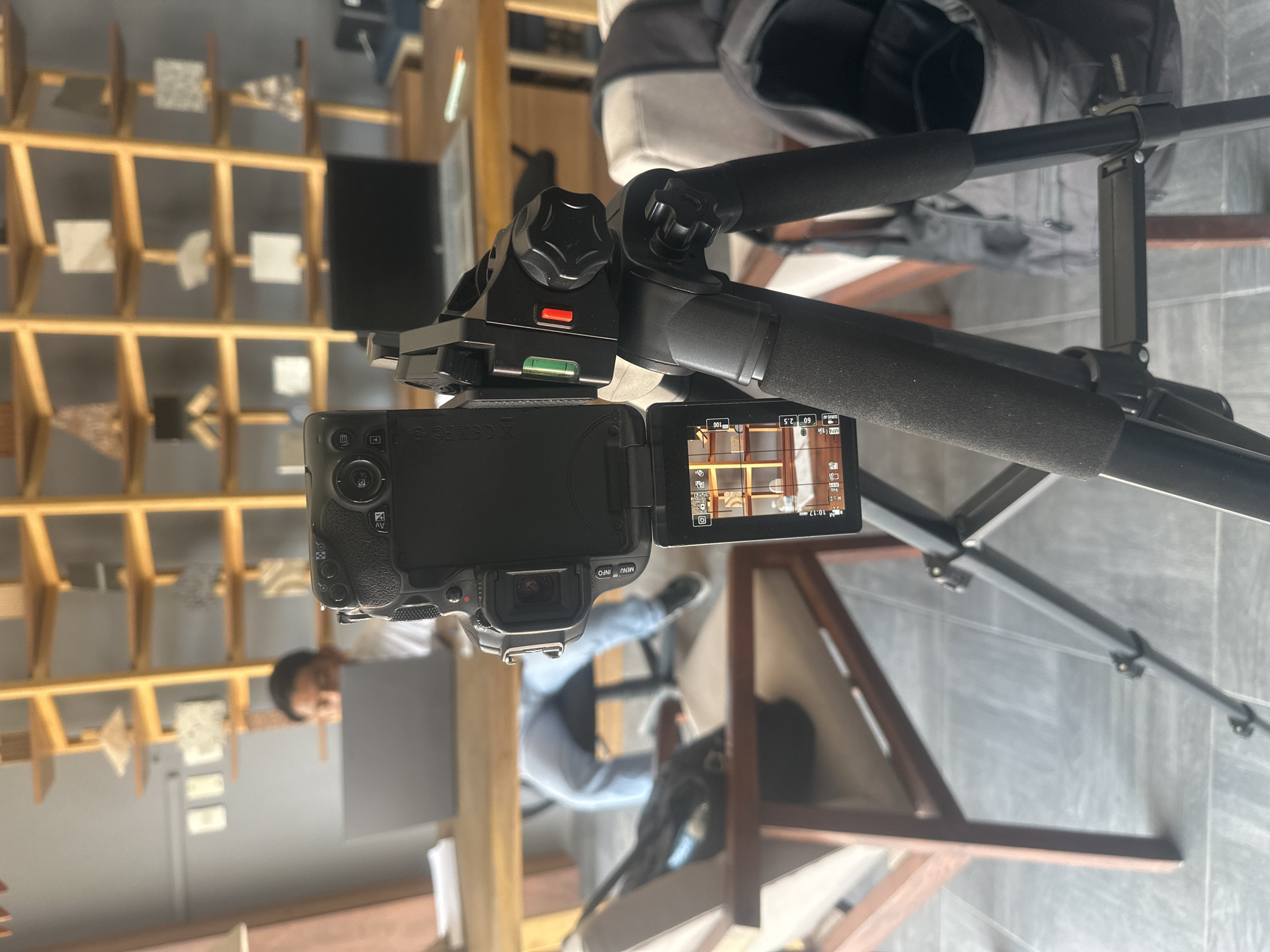 A digital camera, likely a mirrorless or DSLR model, is mounted on a tripod, displaying an image on its articulated screen. The image shown on the screen indicates a view of shelves filled with various objects. The tripod is black with foam grips on the legs.
In the background, shelves are visible, filled with an assortment of tile or stone samples, suggesting a store or showroom for building materials. A person, partially visible, appears to be holding a gray square and looking toward the camera or the scene being filmed. Another person, dressed in a blue shirt, is visible in the background.
The setting might be located in Puebla, Mexico, due to the architectural style and building materials shown.