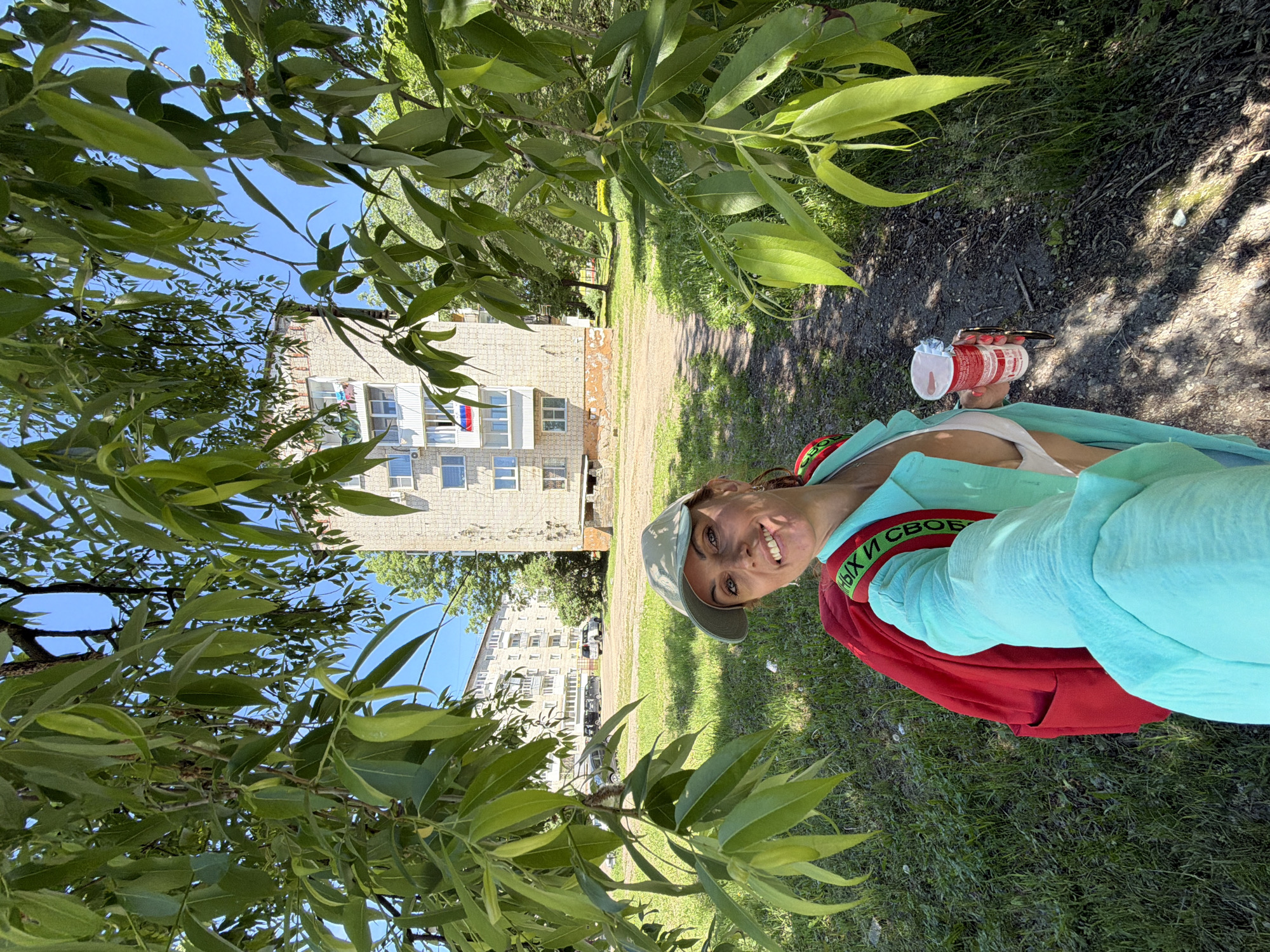 A selfie shows a smiling woman with a red backpack and a light blue shirt, wearing a baseball cap. She holds a cup in her right hand, and sunglasses in her left. Behind her, a tree branches out, partially obscuring a brick apartment building with a Russian flag on a balcony. The background includes grass, a dirt path, and other residential buildings typical of Birobidzhan, Russia.