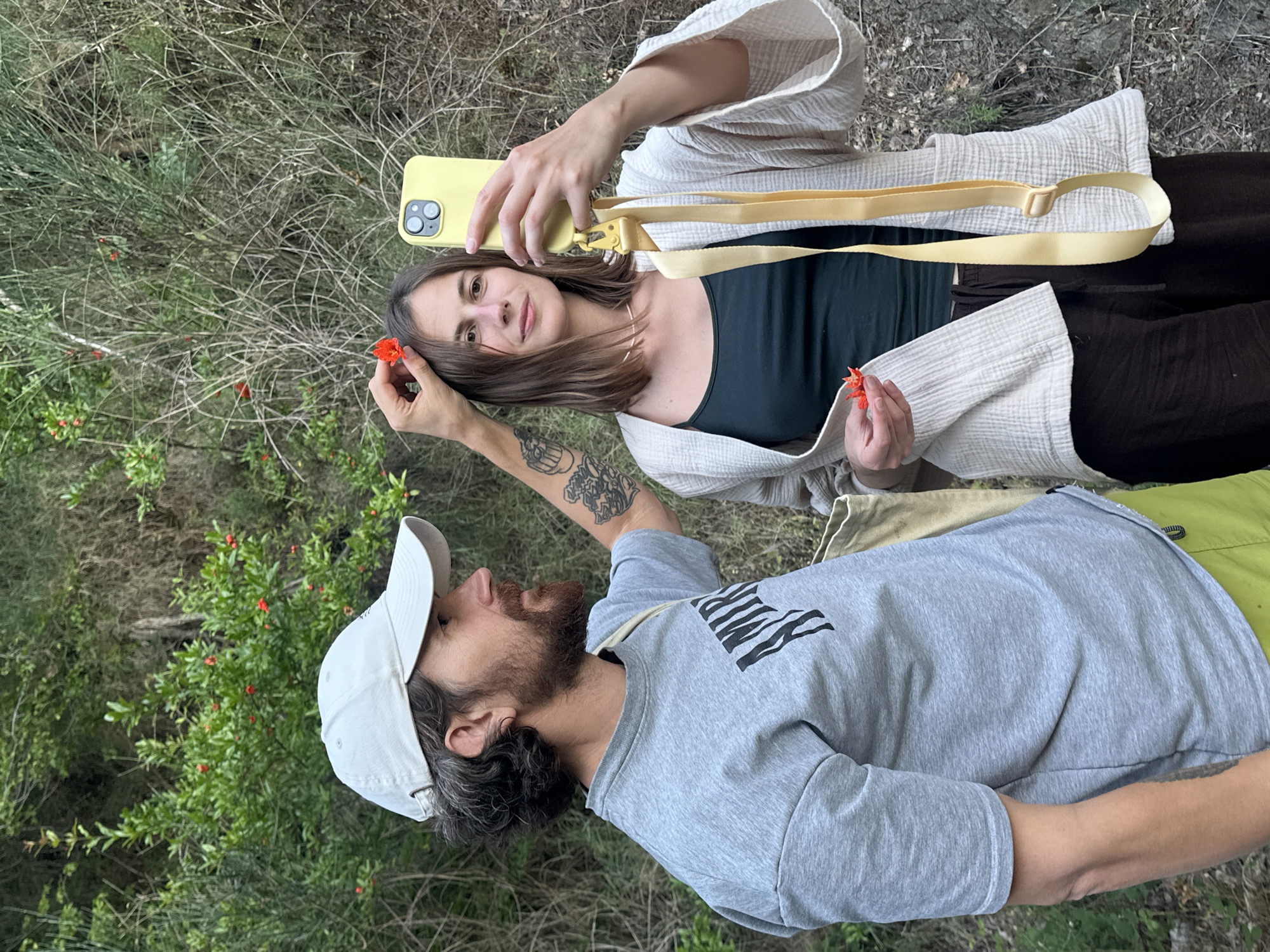 The image shows two people in an outdoor, natural environment, possibly a wooded area or hillside, with dense green foliage and dry branches in the background. A man wearing a light-colored baseball cap, a grey t-shirt with black lettering, and green shorts stands on the left. He has a beard and tattoos on his arm. A woman is on the right, wearing a dark top and a light-colored, open-fronted jacket with wide sleeves. She holds a yellow smartphone in her right hand, seemingly taking a selfie, and has a bright red flower in her left hand. The man is reaching towards her head with his left hand, holding another red flower. The overall impression is a casual, candid moment shared between the two individuals amidst nature. The specific location in Rafailovići, Montenegro cannot be determined from the image alone.