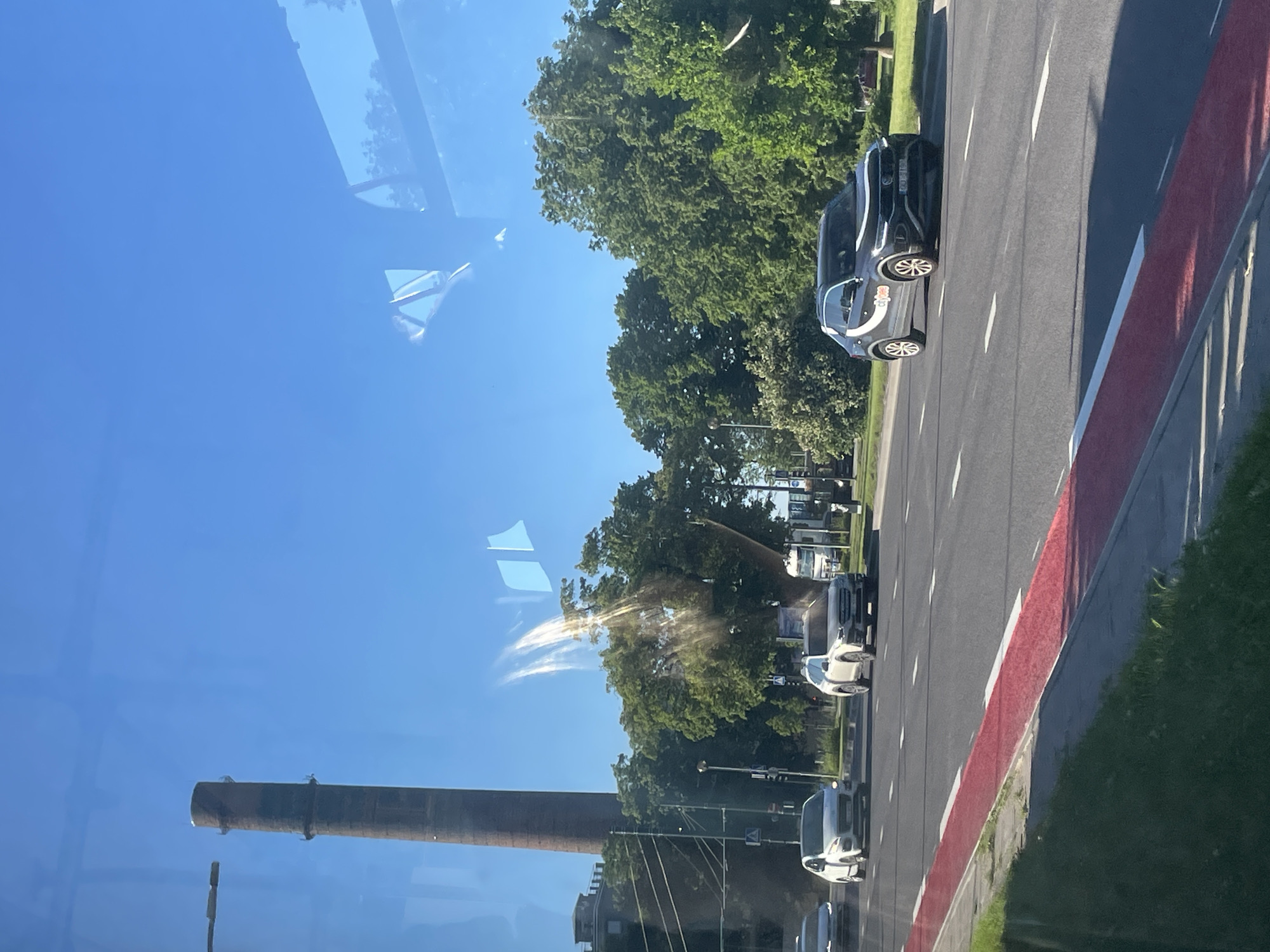An eye-level, medium shot captures a sunny daytime scene on a multi-lane road in Tallinn, Estonia, viewed from inside a vehicle. A tall, reddish-brown industrial smokestack, likely a relic of the past, dominates the left side of the frame, piercing a clear blue sky. Beneath the smokestack, lush green trees form a dense canopy. The road features white lane markings and a distinct red-painted lane, possibly a bike lane or a specially designated zone. Several cars are visible on the road: a white Audi SUV is positioned in the right lane, while a white hatchback and a lighter-colored car are further ahead in the left lanes. A dark gray Volkswagen T-Roc SUV, with white decals on its side, is in the right lane, ahead of the Audi. Reflections of the sky and trees are visible on the vehicles and the glass through which the photo is taken, suggesting the image was captured through a window. The overall impression is one of urban infrastructure meeting natural elements under a bright, clear sky.