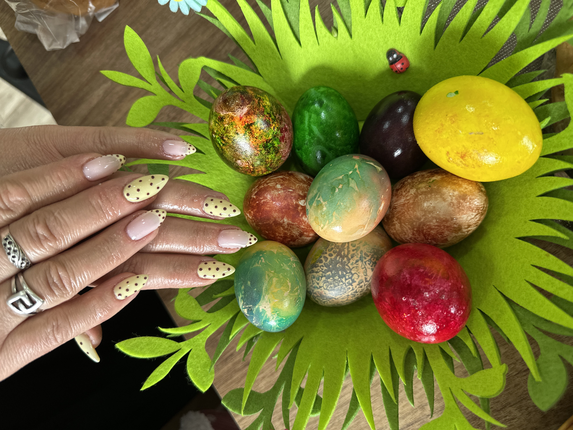 The scene shows a close-up of a hand with decorated nails holding a green felt basket filled with nine colorful Easter eggs. The eggs vary in color, including yellow, brown, green, red, and blue. A small ladybug decoration is visible on the basket. The nails are decorated with a light pink base and yellow tips with brown polka dots. The hand is also adorned with two silver rings. The background appears to be a wooden surface. The image was taken in Lehchevo, Bulgaria.