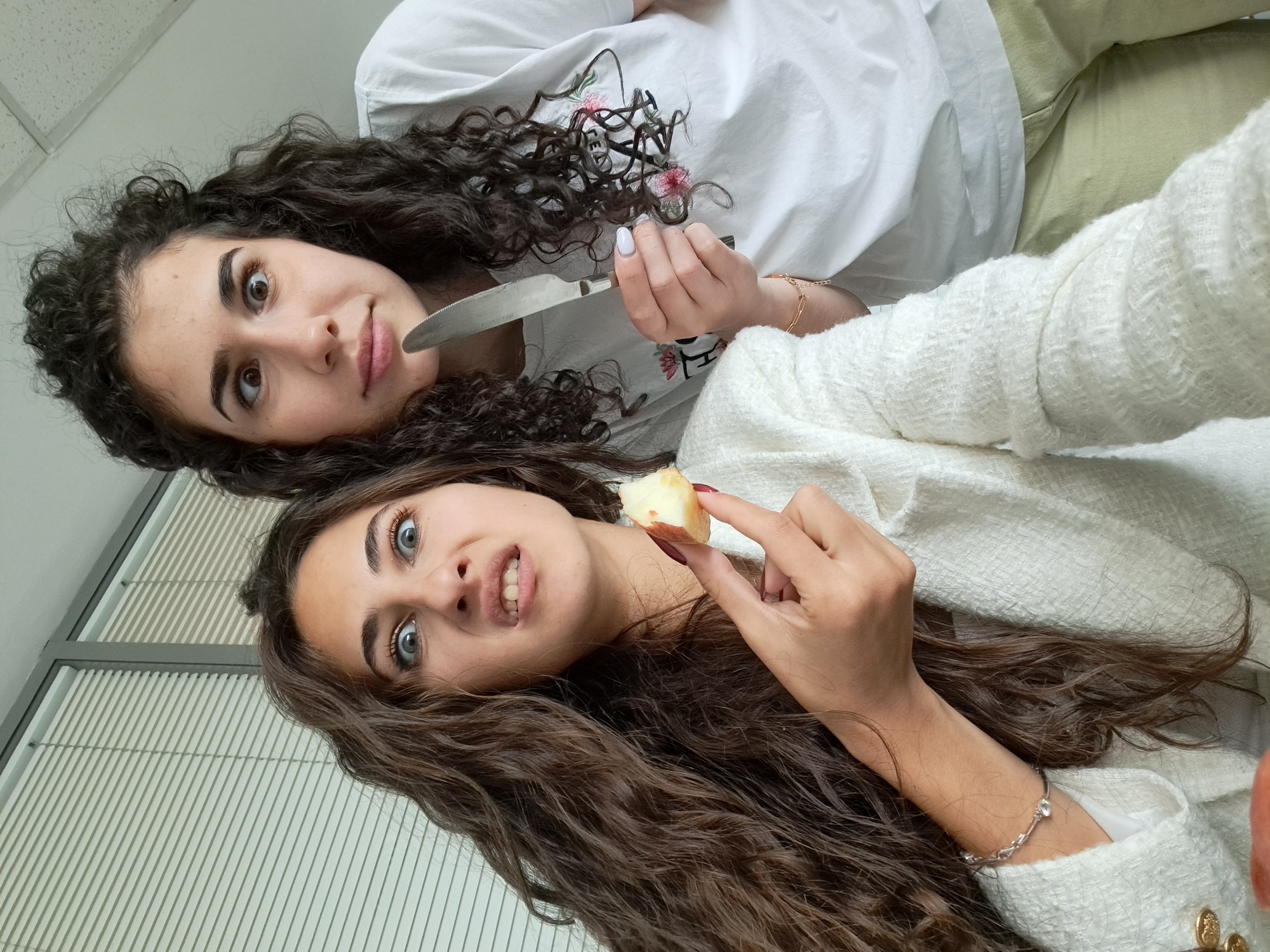 Two women pose for a selfie, oriented at an angle. The woman on the left has dark, curly hair, light skin, and is wearing a white shirt with a floral design. She is holding a knife. The woman on the right has long, wavy brown hair, light skin, and is wearing a white jacket. She is holding a piece of fruit. The background includes a white ceiling and a window with closed blinds. It is not possible to identify the exact location in Sofia, Bulgaria.