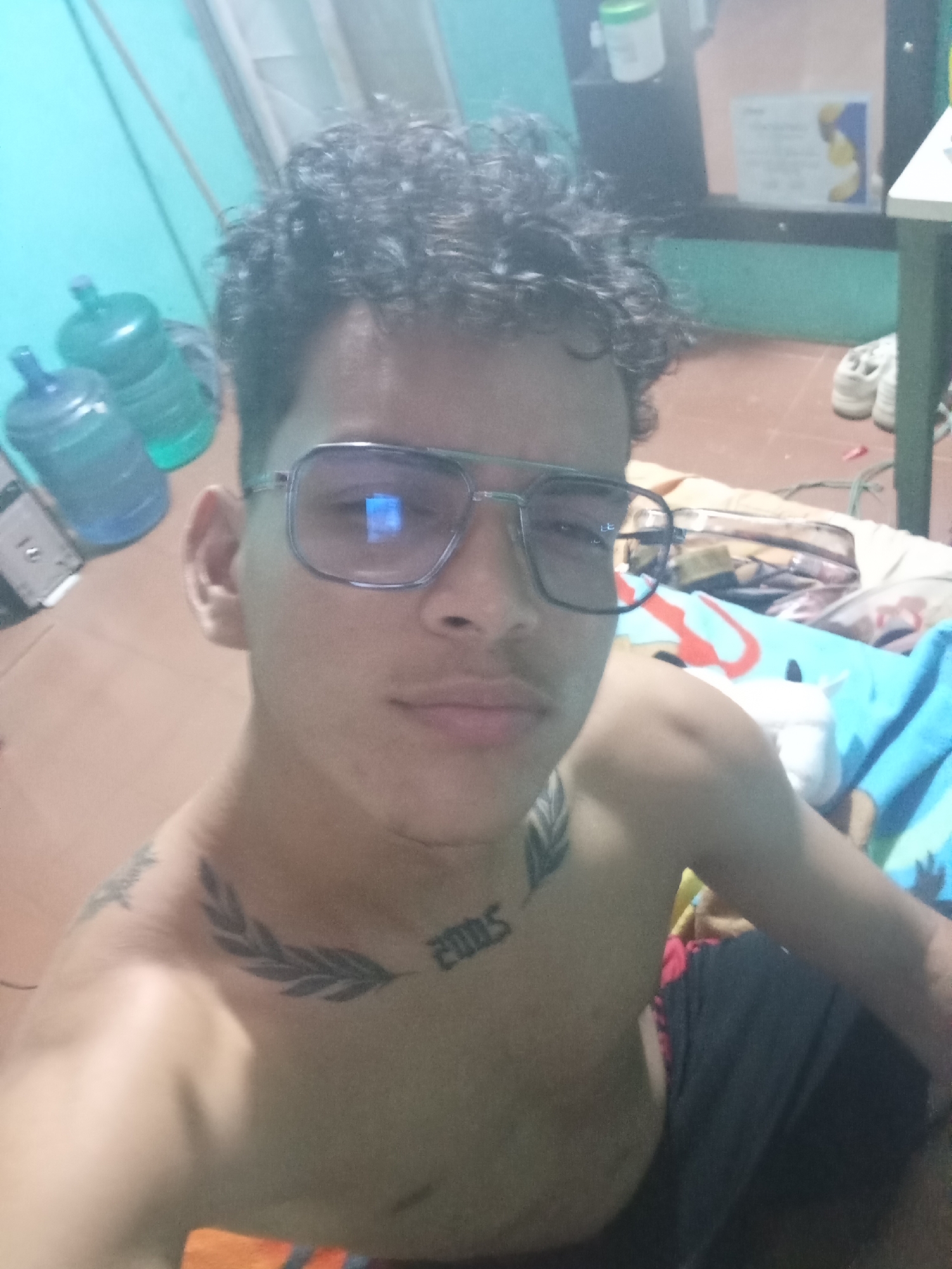 A shirtless young man with dark, curly hair and glasses poses for a selfie indoors. He has tattoos on his chest and shoulder. The background shows a turquoise-colored wall, water jugs, and a cluttered area with various objects, including bedding and shoes. The location in the city of Altagracia, Venezuela is not discernable from the image.