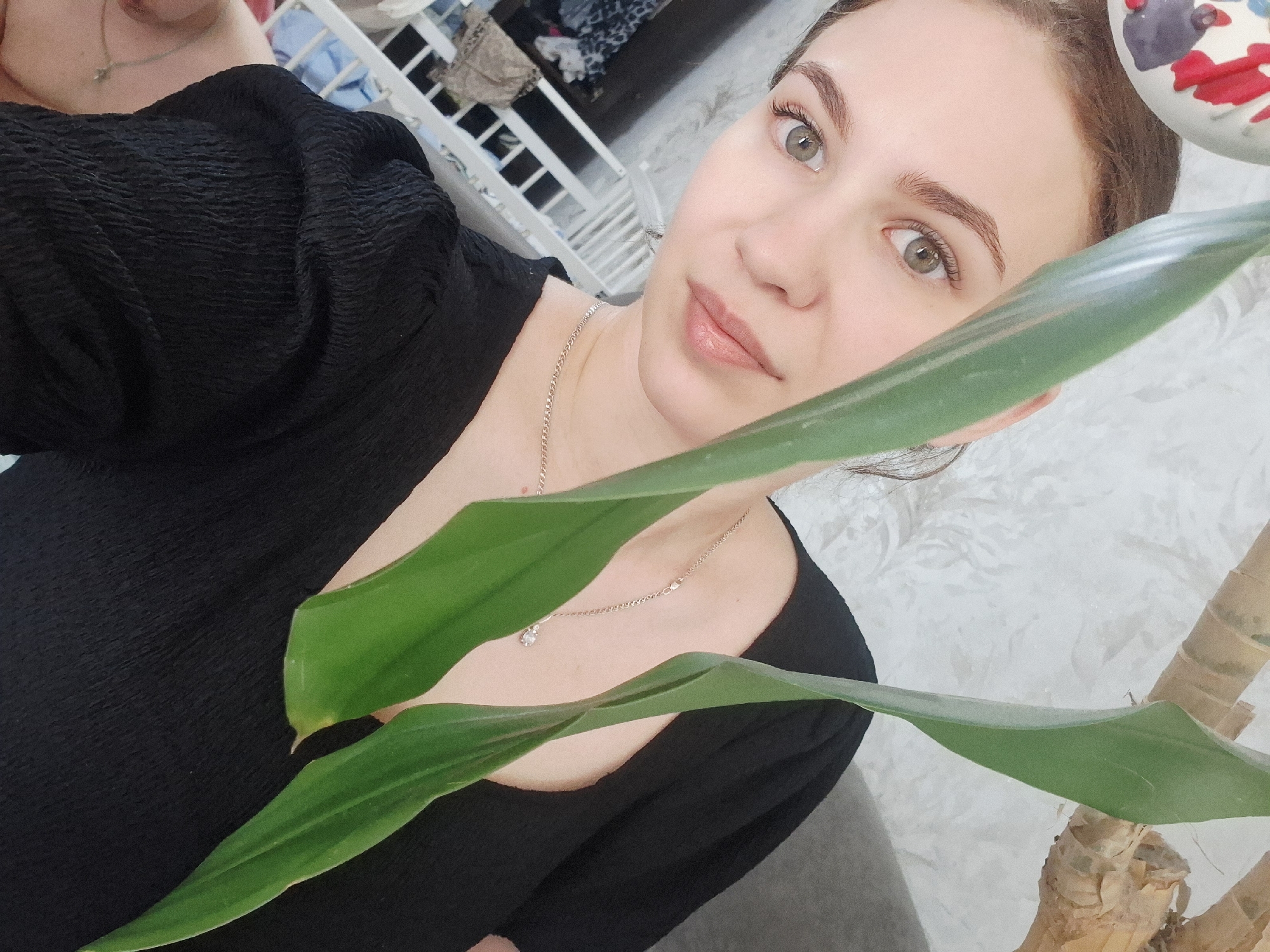 A woman with light skin and brown hair, wearing a black blouse and a silver necklace with a pendant, takes a selfie. She is indoors, as indicated by the patterned wallpaper in the background and other interior elements like a white crib. A potted plant with large green leaves partially obscures the view. The location is Chaikovsky, Russia.