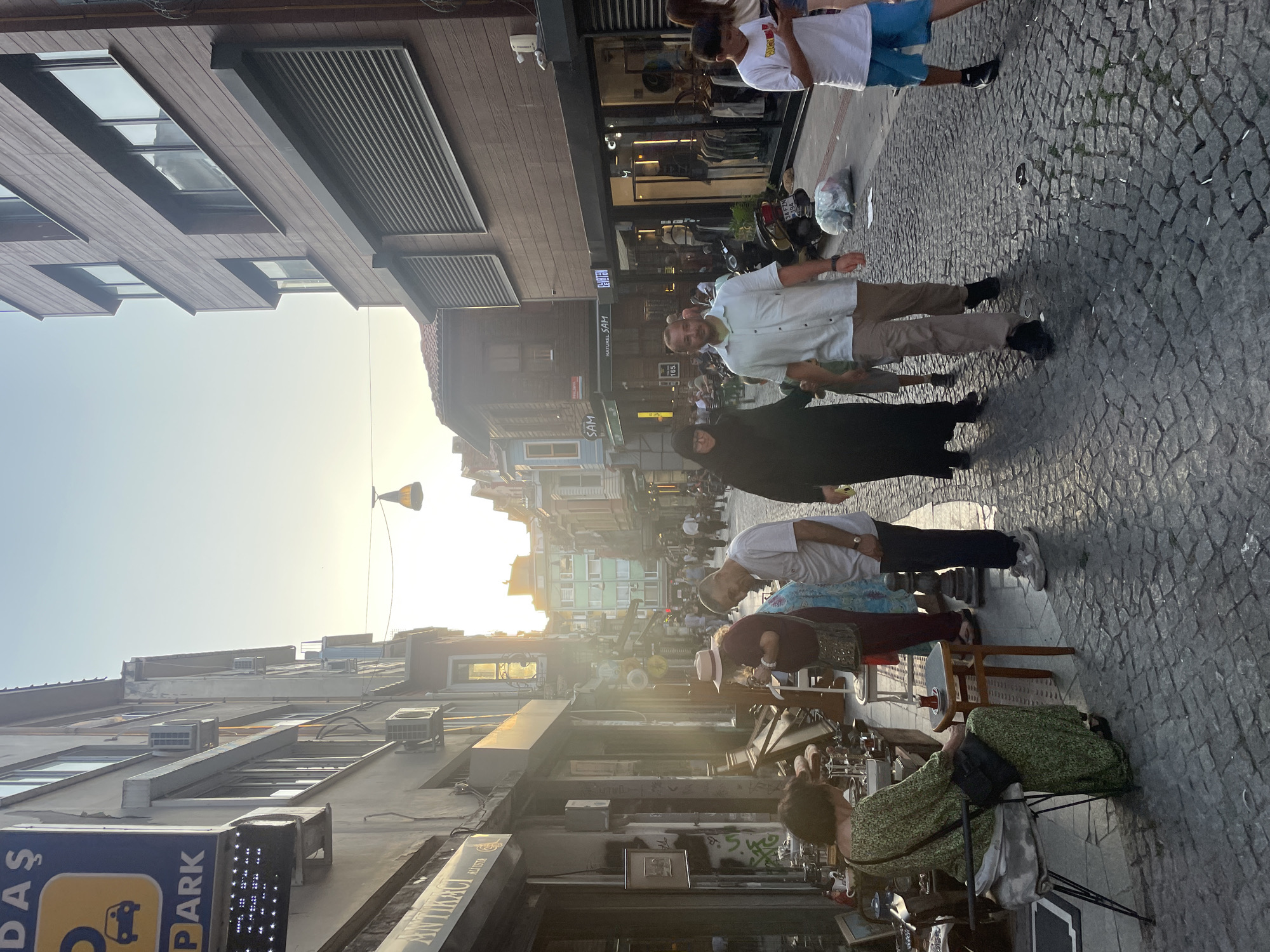 The image depicts a cobblestone street scene in Kadıköy, Turkey, bathed in the warm glow of late afternoon sunlight. The street is lined with multi-story buildings, some with traditional architecture and others with more modern facades featuring wooden paneling and metal shutters. Shops are visible on the ground floor, with one storefront displaying antiques and curios. Several people are walking along the street and interacting. In the foreground, a woman in a green dress sits on a stool near an antique stall. A couple, a man in a white shirt and a woman in a black abaya, walk together. Further down the street, a group of people are browsing or walking. The overall atmosphere suggests a bustling commercial and residential area.