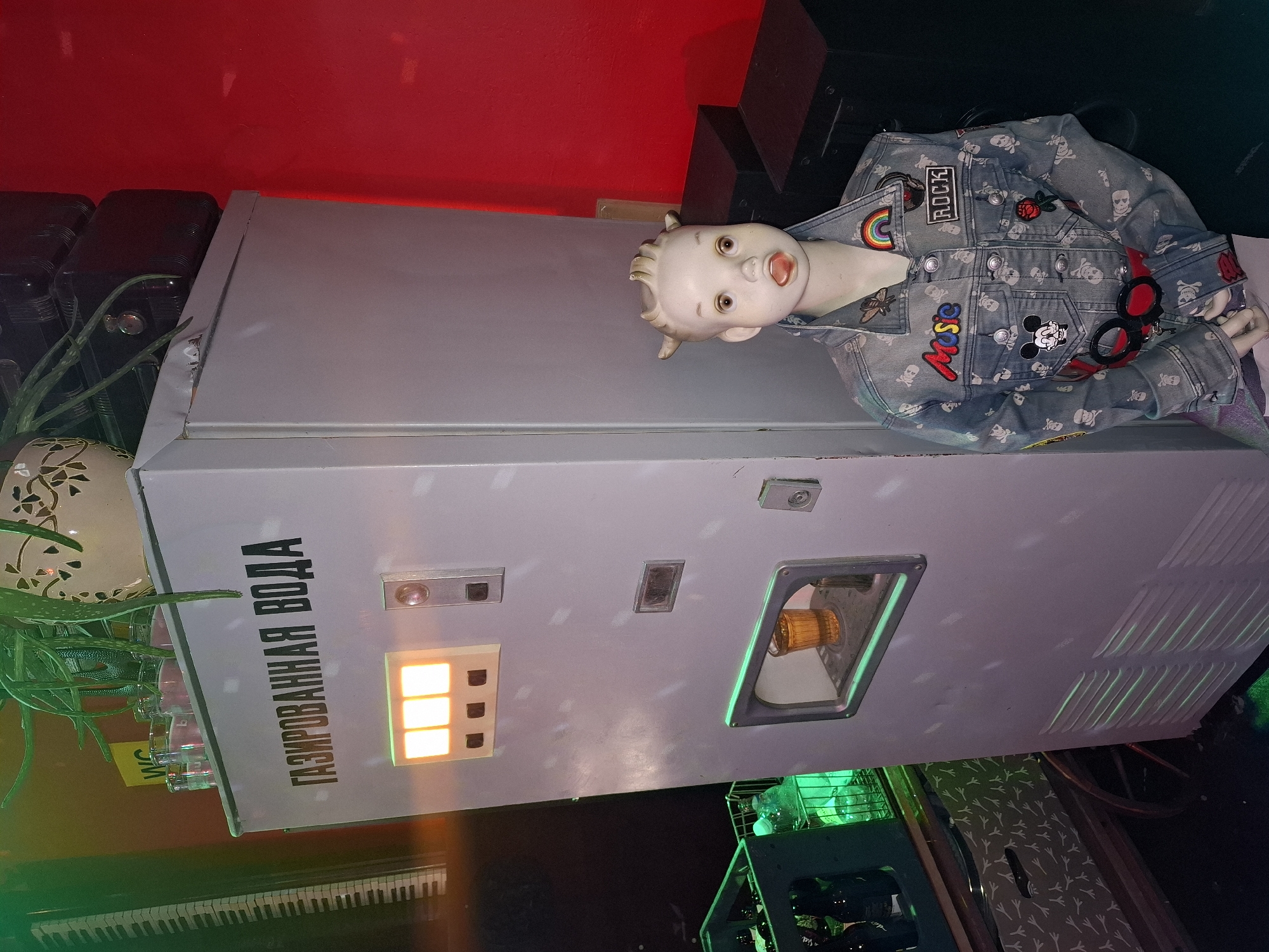 The image presents a quirky and surreal scene. A vintage-style, light-gray soda dispenser with the inscription "ГАЗИРОВАННАЯ ВОДА" (carbonated water) in Russian is the central element. The dispenser features illuminated orange lights and a dispensing outlet. Resting on top of the dispenser is a porcelain-like doll figure, wearing a denim jacket adorned with patches and buttons. The background includes a red wall, various items suggesting a bar or entertainment setting, and what appears to be a keyboard. The location is likely a bar or club in Riga, Latvia, based on the Russian inscription on the soda dispenser.