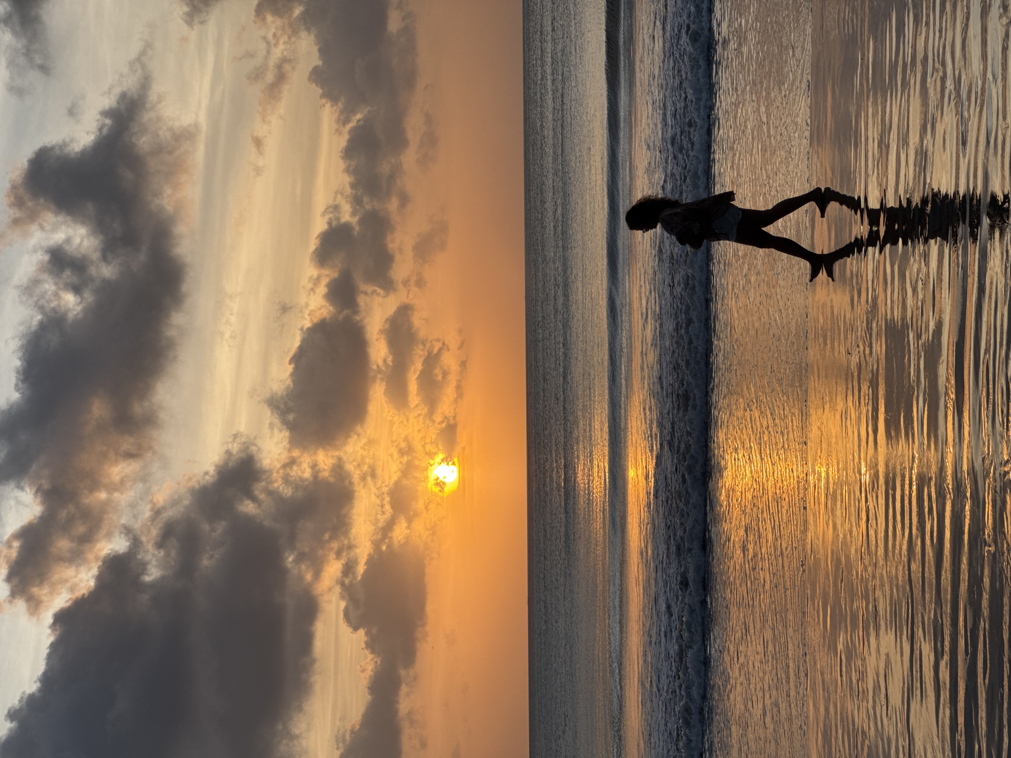 The image is divided into two vertical panels. The left panel depicts a sky at sunset with orange hues, dark clouds, and the sun visible through the clouds. The right panel shows a beach scene with a person standing in the shallow water. The sunset's reflection is visible on the wet sand. The silhouette of the person is also reflected. The scene likely takes place in the city of Kuta Selatan, Indonesia.
