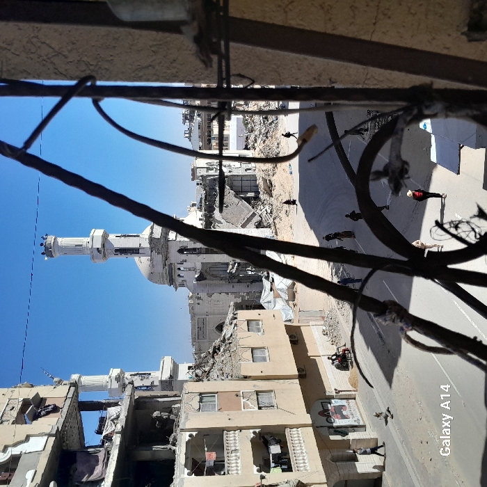 The image depicts a street scene in Khan Yunis, Palestinian Territory, characterized by extensive structural damage under a clear blue sky. The foreground is partially obscured by dark, rusty metal bars, suggesting the photo was taken through a damaged window or balcony railing.
In the midground, a paved road stretches through the urban area. Approximately fifteen individuals are visible on the street and sidewalks, walking or standing. On the left, a multi-story building shows severe damage, with upper floors partially collapsed, exposing rebar and interiors. Laundry hangs from balconies on its lower, less damaged sections. Near its base, two people are seated on a bench beside a pile of sand.
Dominating the background is a large mosque with a prominent white dome and a tall, intact minaret featuring crenellated battlements. The mosque itself exhibits significant damage, with large sections of its walls missing and rubble visible around its base, indicating structural compromise. To the right of the mosque, more buildings are visible, also heavily damaged and reduced to rubble in many areas. Piles of debris and a few white tarpaulins are scattered across the scene. The overall environment conveys widespread destruction. The watermark "Galaxy A14" is present in the lower-left corner.
