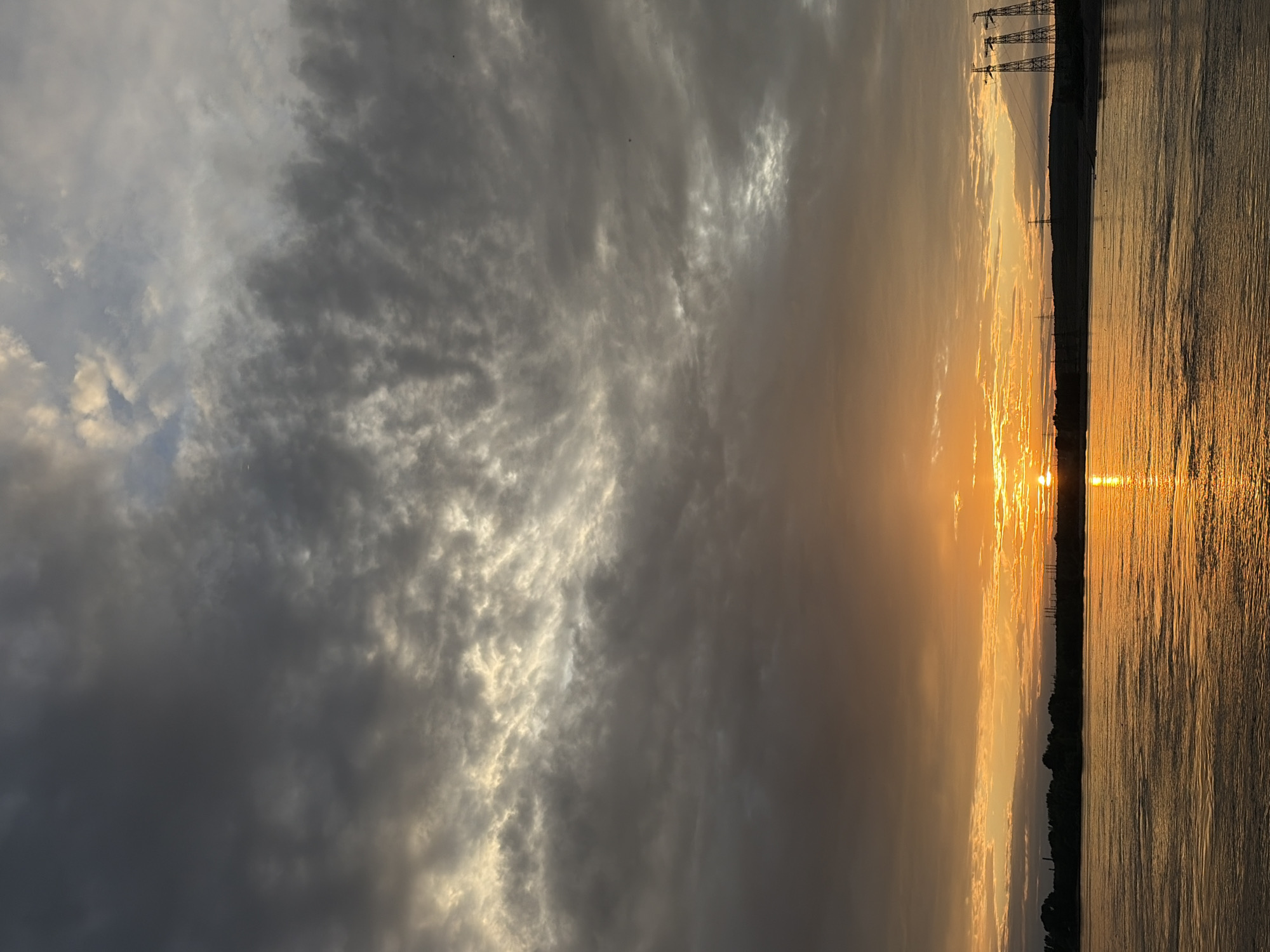 This media file depicts a scenic sunset over a body of water, likely the Kama River, near Chaikovsky, Russia. The scene is dominated by a dramatic sky filled with a mix of dark, heavy clouds and streaks of bright, golden sunlight. The sun itself is low on the horizon, casting a brilliant orange and yellow glow across the sky and reflecting intensely on the rippling surface of the water.
On the far side of the water, a silhouetted treeline is visible, with several tall, lattice-like structures resembling power line pylons interspersed along the horizon. These structures, along with the dense clouds, suggest an industrial or semi-industrial landscape bordering the natural beauty of the sunset. The overall mood is one of awe and tranquility, with the dramatic weather adding to the visual spectacle. The time of day is clearly sunset, indicated by the low angle of the sun and the warm, rich colors of the sky and water. No people are visible, and there is no visible text in the image.