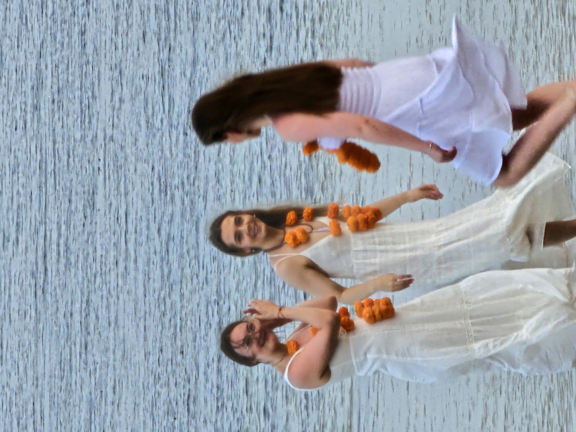 In Denpasar, Indonesia, three women wearing white dresses and orange marigold garlands stand in shallow water, possibly a beach or riverbank. The woman on the left, with glasses and pulled-back hair, is facing left and appears to be adjusting her hair with her hands. The woman in the center, with long brown hair, is looking forward and smiling. The woman on the right, also with long brown hair, is facing away from the camera and her white dress is billowing around her legs. The water is a muted blue-grey with ripples, suggesting a calm body of water. The lighting is soft and diffused, indicating it may be overcast or late afternoon.