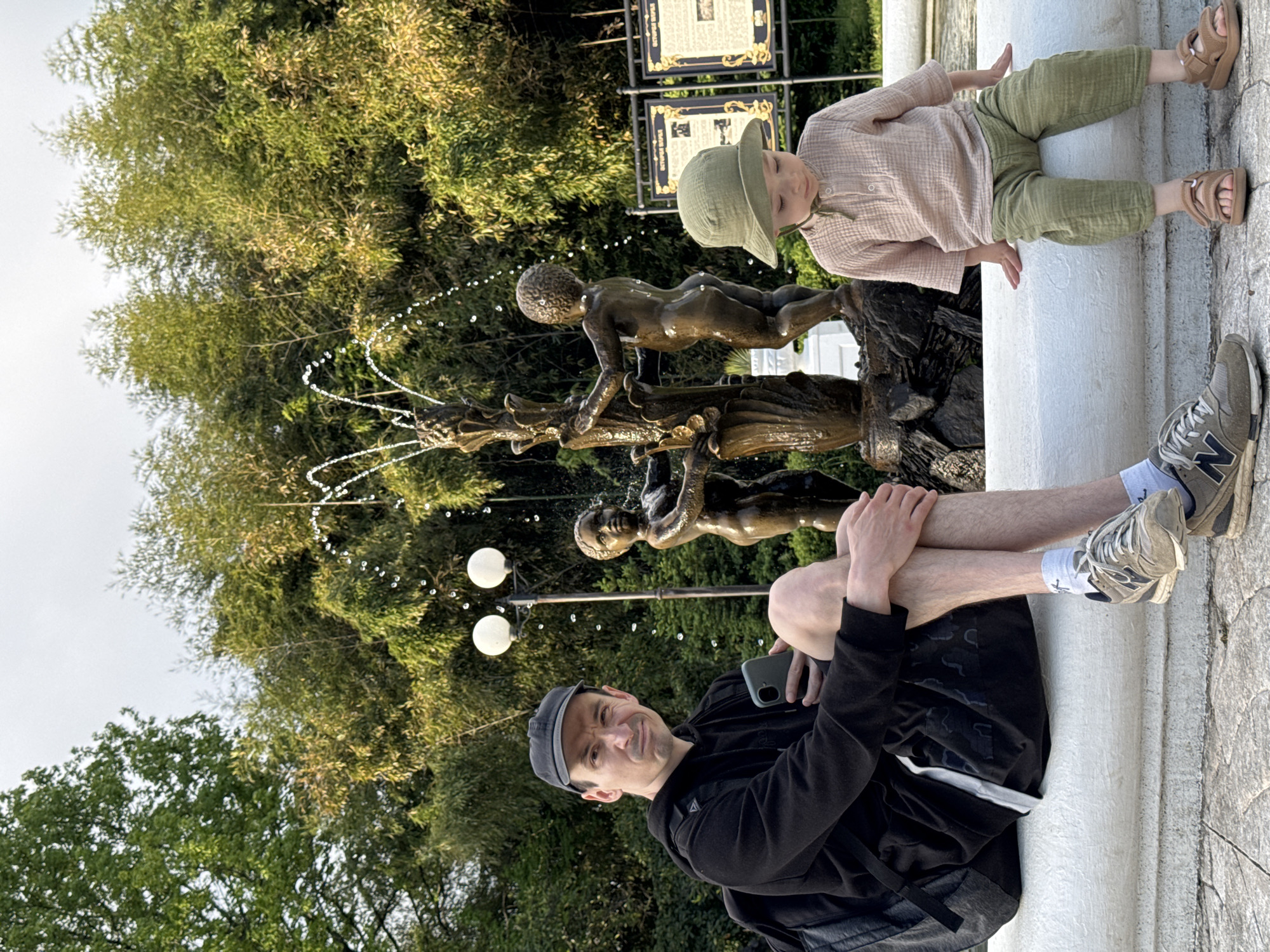 A father and young child are sitting along the edge of a fountain in a park. The fountain features bronze statues of children. The father is seated with his legs bent at the knees, and appears to be looking at his phone. The child is standing next to the fountain, appearing to interact with the statues. The location is in Sirius, Russia. The background includes trees and what appear to be informational signs.