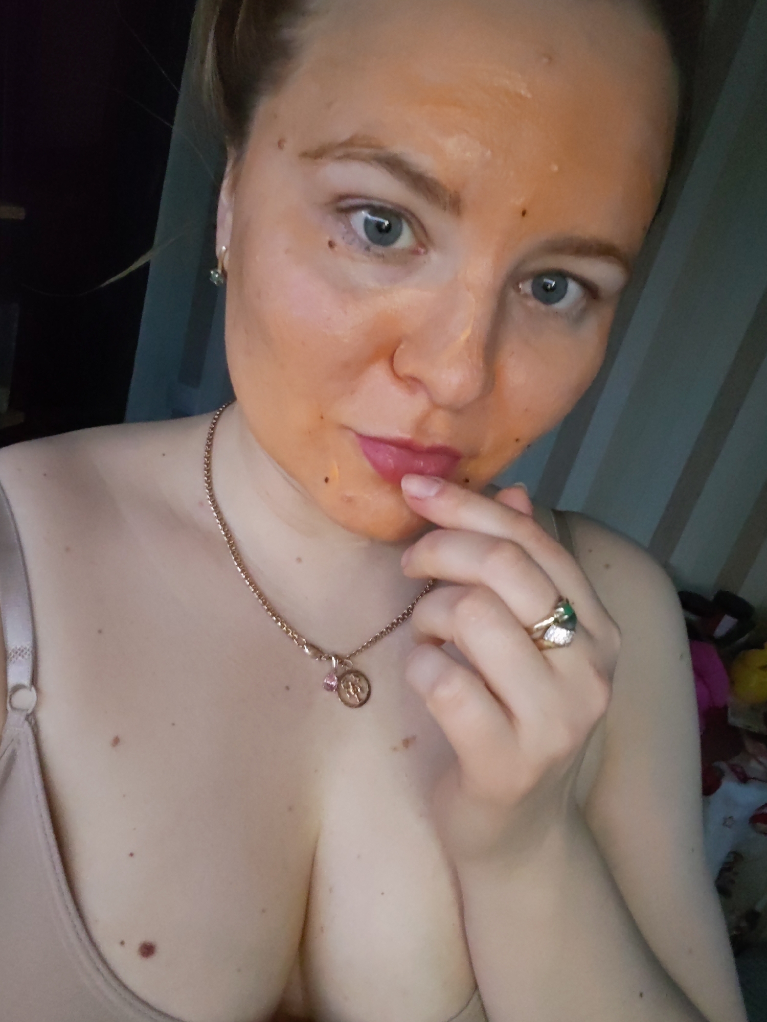 A light-skinned female with an orange face mask is posing for a selfie. She has blue eyes and light-colored hair pulled back. She is wearing a beige bra, a gold necklace with a pendant, and a gold ring with green and clear stones. She is holding her finger to her lips. The background is blurry, but includes vertical stripes and possibly bedding. There is no visible evidence to confirm the location in Izhevsk, Russia.
