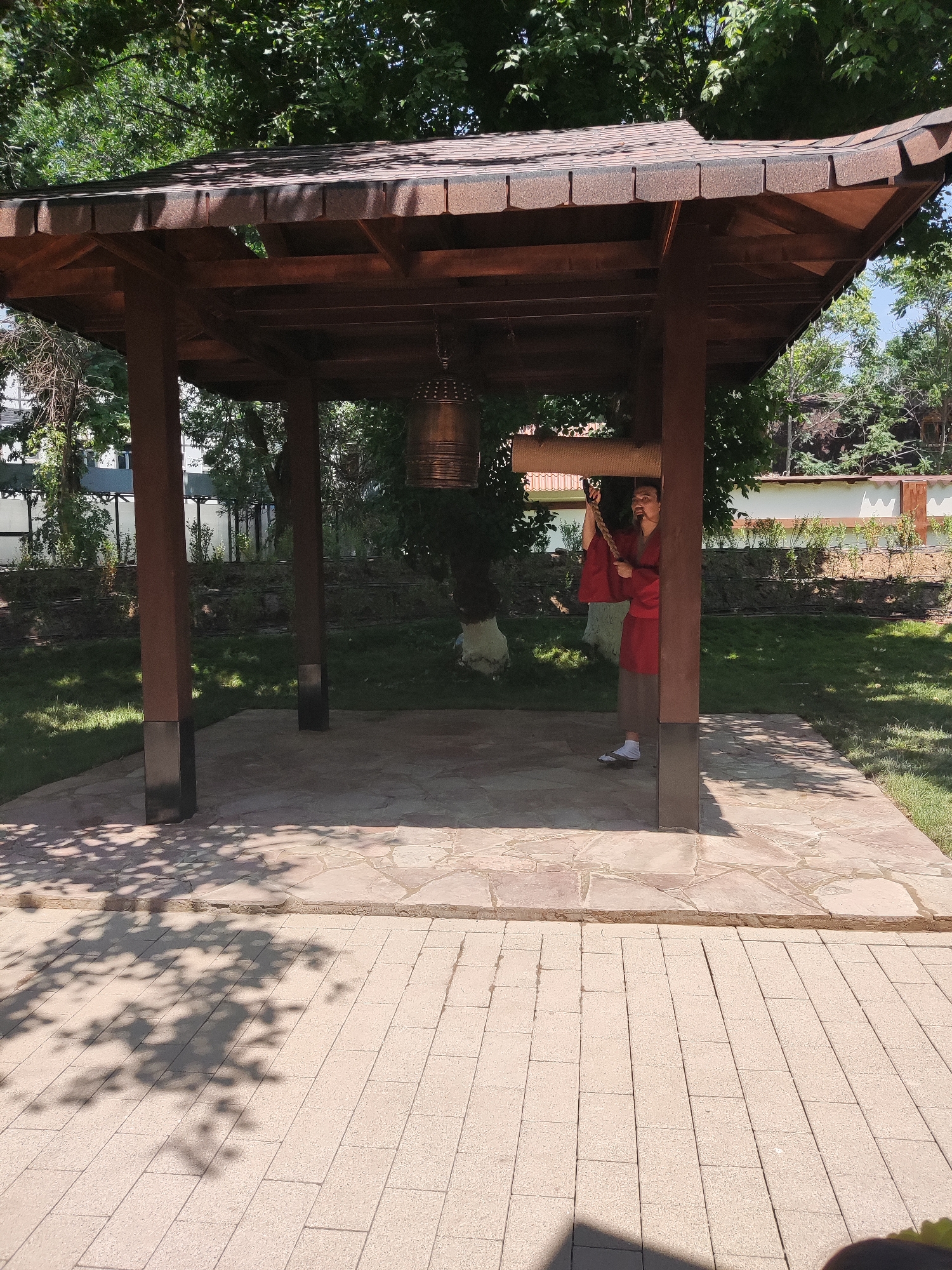 A person wearing a red robe and brown pants stands inside a wooden gazebo with a large bell hanging from the roof. The person is holding a rope attached to the bell, appearing to be about to ring it. The gazebo has a brown tiled roof and dark brown support posts. The ground inside the gazebo is paved with irregular flagstones, and the area outside is covered with light-colored rectangular paving stones. Green grass surrounds the gazebo, with trees and a fence visible in the background. The scene is bathed in sunlight, casting dappled shadows on the ground. This location appears to be within the Botanical Garden in Shymkent, Kazakhstan, a popular urban green space.