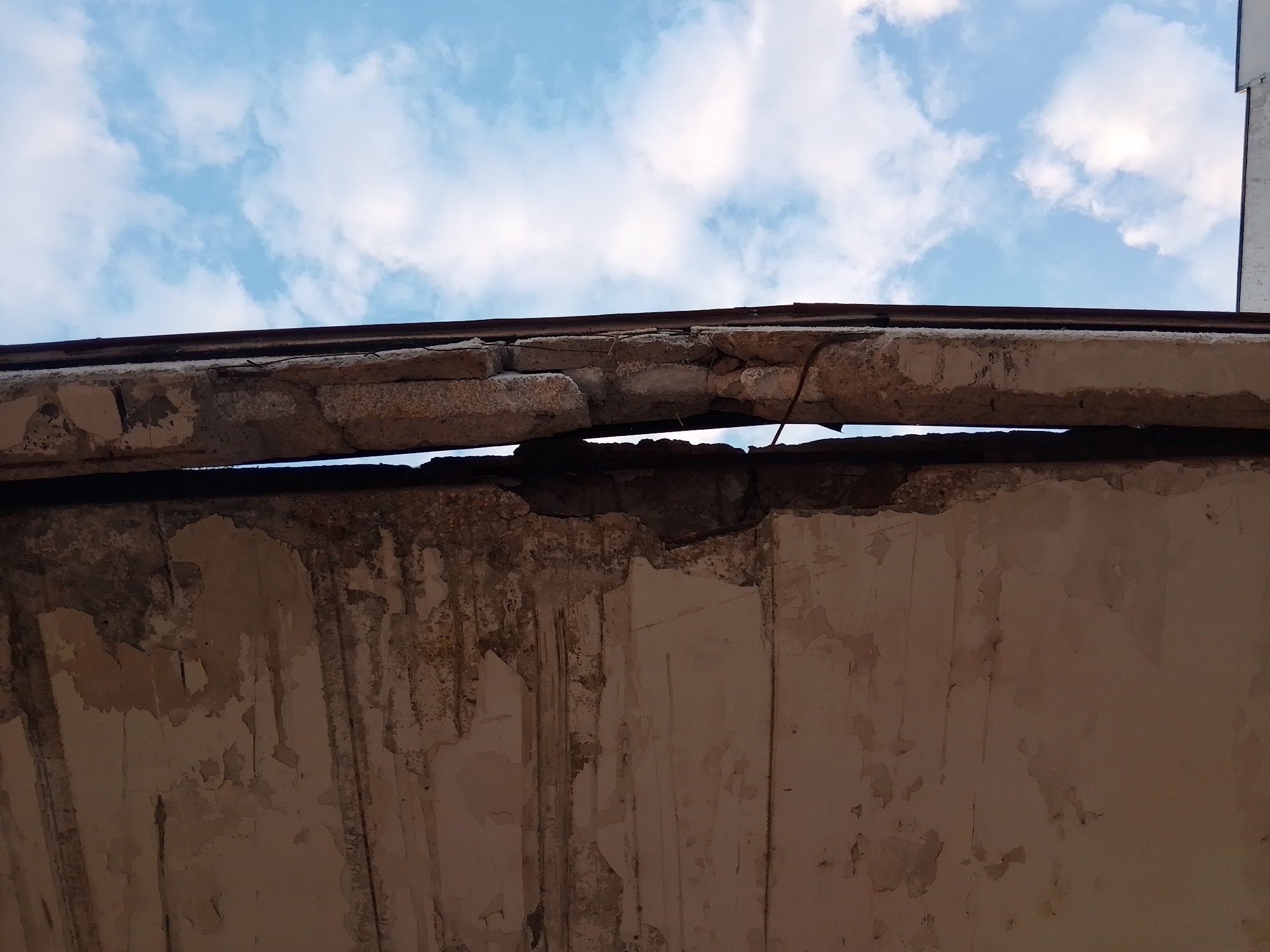 The image captures a severely deteriorated structural element of a building, viewed from below against a partly cloudy blue sky. The upper section reveals a heavily damaged concrete beam or lintel, fractured into multiple segments with significant spalling and exposed rebar, particularly on the right side. A thin, dark linear element rests directly above this damaged beam. Below the beam, a wall surface exhibits extensive signs of decay, including widespread peeling paint or plaster, dark vertical streaks indicative of water damage or staining, and general discoloration. A visible gap exists between the damaged upper beam and the wall below. The scene conveys significant structural compromise and long-term neglect in the city of Unknown, Unknown. No individuals, animals, or discernible actions are present.