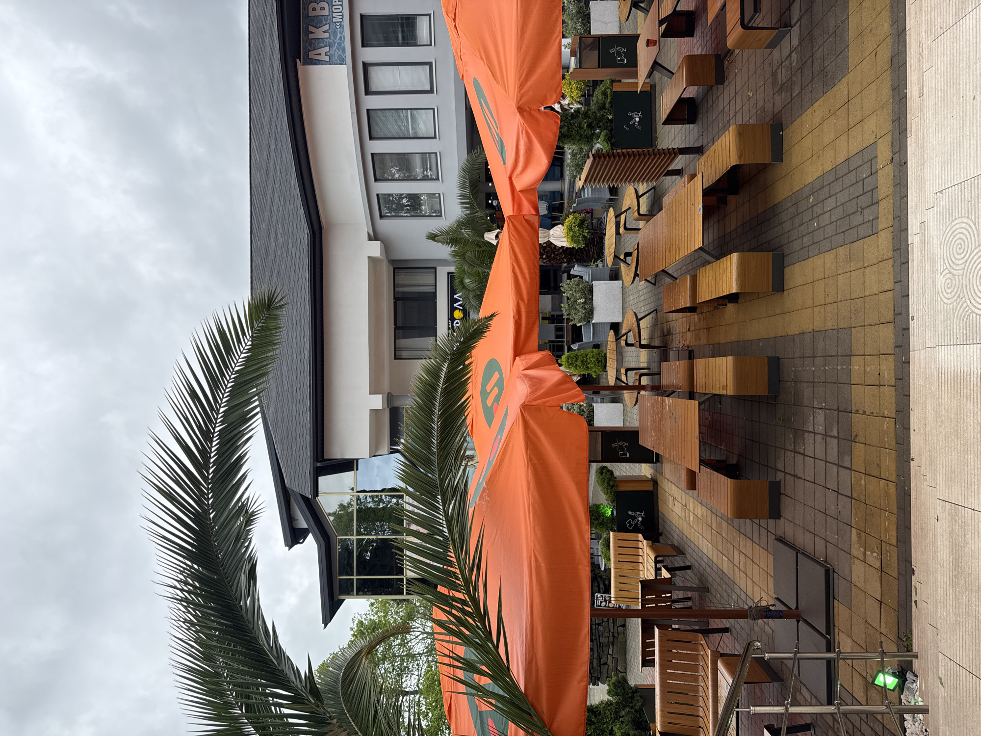 The image depicts an outdoor café setting, likely in Sochi, Russia. The location appears to be an urban area with a combination of modern architecture and tropical elements, suggested by the presence of palm trees. The café furniture consists of wooden tables and benches. The tables are arranged on a paved walkway constructed from yellow and grey bricks. Large orange canopies with a logo provide shade over some tables.
There is a multi-story building with a grey roof and white walls. The sign "AKB" is visible on the building. The weather appears to be overcast or rainy, as the pavement is wet. There are no people visible in the image. There are plant boxes behind the tables.