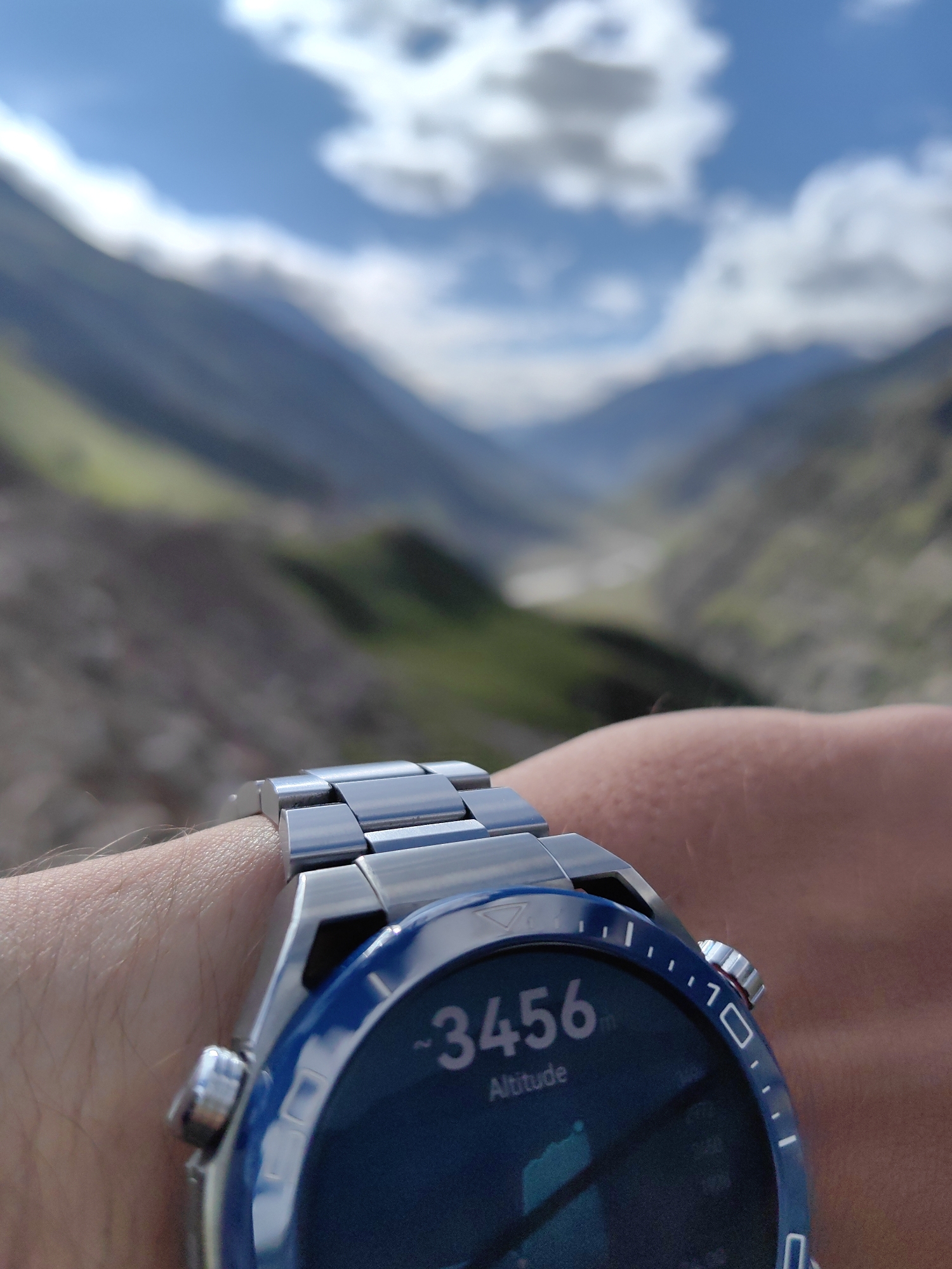 An outdoor, close-up, eye-level shot shows a smartwatch on a person's wrist. The smartwatch has a blue bezel and a digital display showing an altitude of 3456m. The watchband is made of polished metal links. The background is out of focus, showing a mountainous landscape with green slopes and a cloudy blue sky. The lighting suggests daylight. The location is unknown, India.