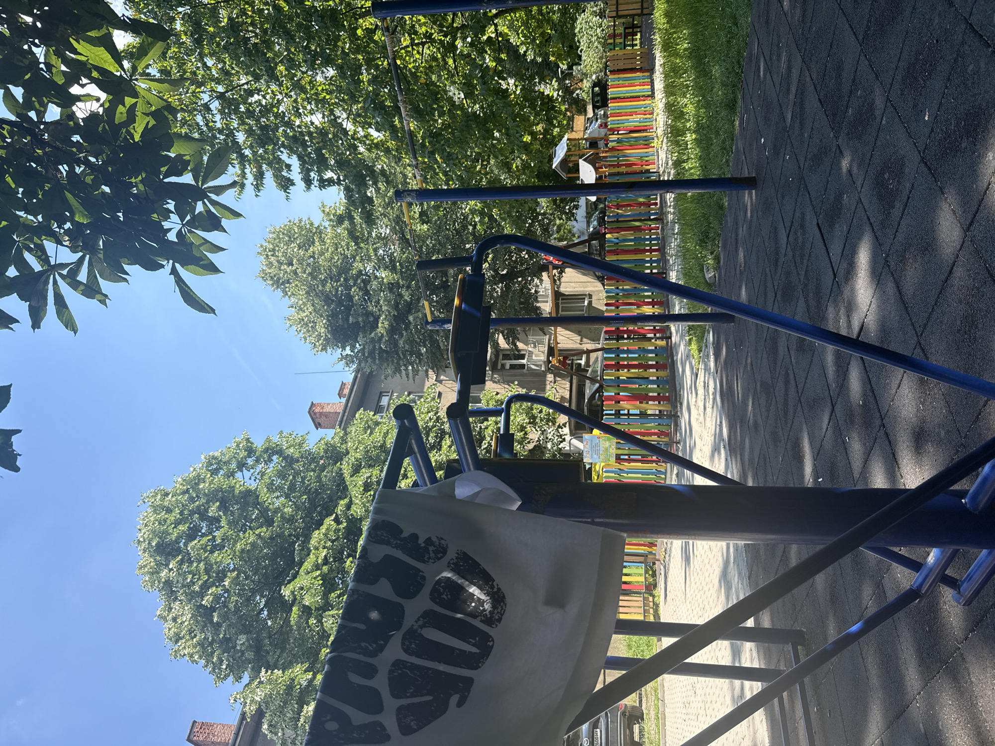 The image shows an outdoor scene on a sunny day. In the foreground, a piece of clothing with black lettering is hanging on a blue metal structure, possibly part of a gym or playground equipment. The lettering on the clothing is partially visible and appears to say "PROTECT YOUR". Behind the metal structure, a colorful fence with vertical red, yellow, blue, and green posts encloses a playground. Beyond the fence, there is a building with red brick chimneys and green trees. The ground in the foreground is covered with dark grey rubber tiles, and there is grass and a paved area visible. A black car is parked on the left side of the image. The sky is clear and blue. The scene appears to be in a residential area, likely a courtyard or park within the city of Sofia, Bulgaria, given the architectural style of the building and the presence of playground and gym equipment in a shared urban space.