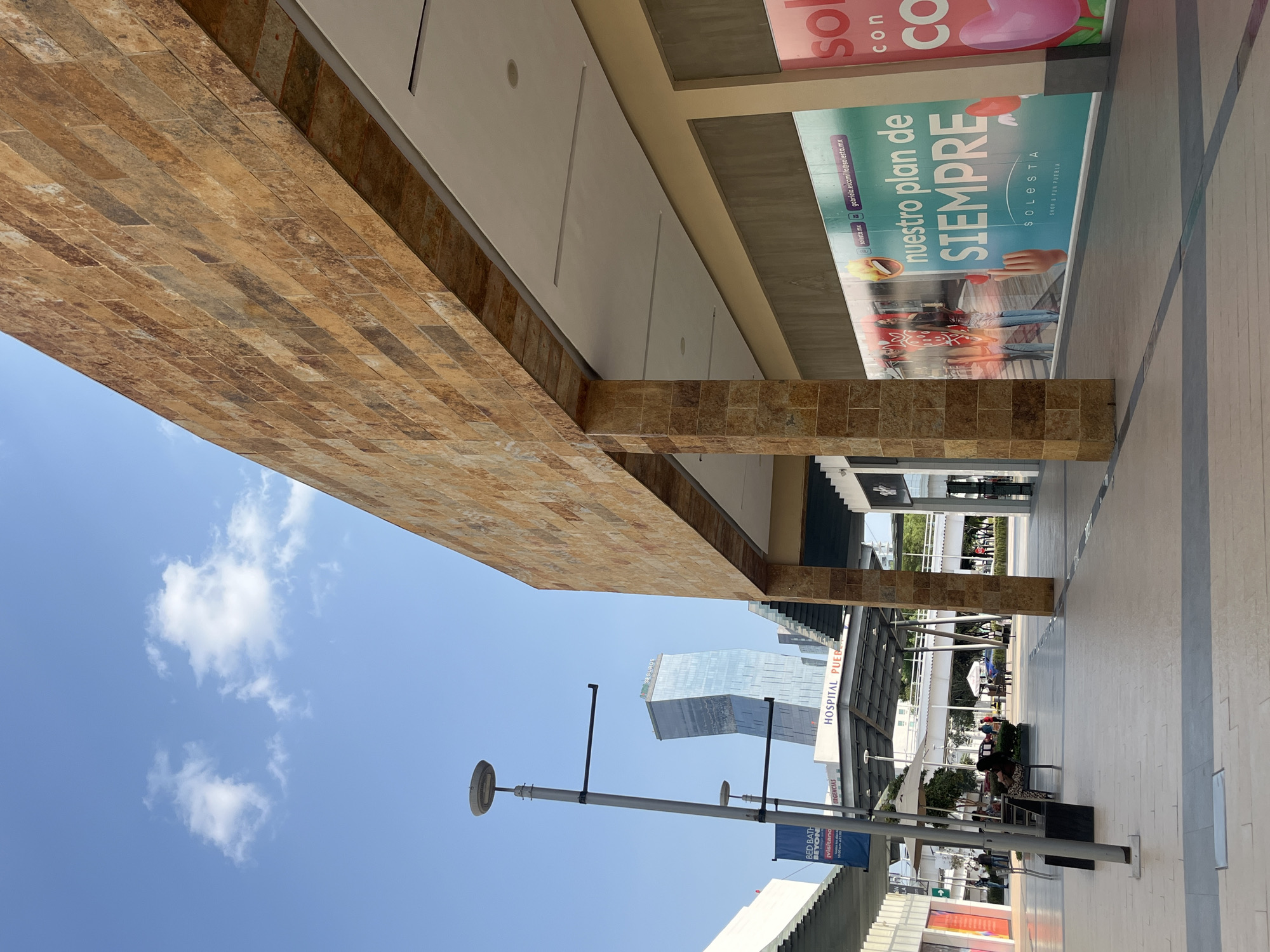 Here is a detailed description of the image:
The image shows an outdoor covered walkway, possibly a part of a shopping center or commercial area. The walkway has a tan-colored, brick-like ceiling that is extended over the walking area with supporting columns made of the same brick material. The walking surface is made of light-colored pavement.
On the right side of the image, there is an advertisement with text in Spanish. The ad features images of people.
In the distance, a tall, modern skyscraper can be seen. Adjacent to it, the sign “HOSPITAL PUEB” is visible. Also visible is a light post and a sign reading “BED BATH BEYOND”
The sky is blue with scattered clouds.
The scene appears to be a well-lit, public space with a combination of architectural elements, commercial signage, and urban infrastructure.