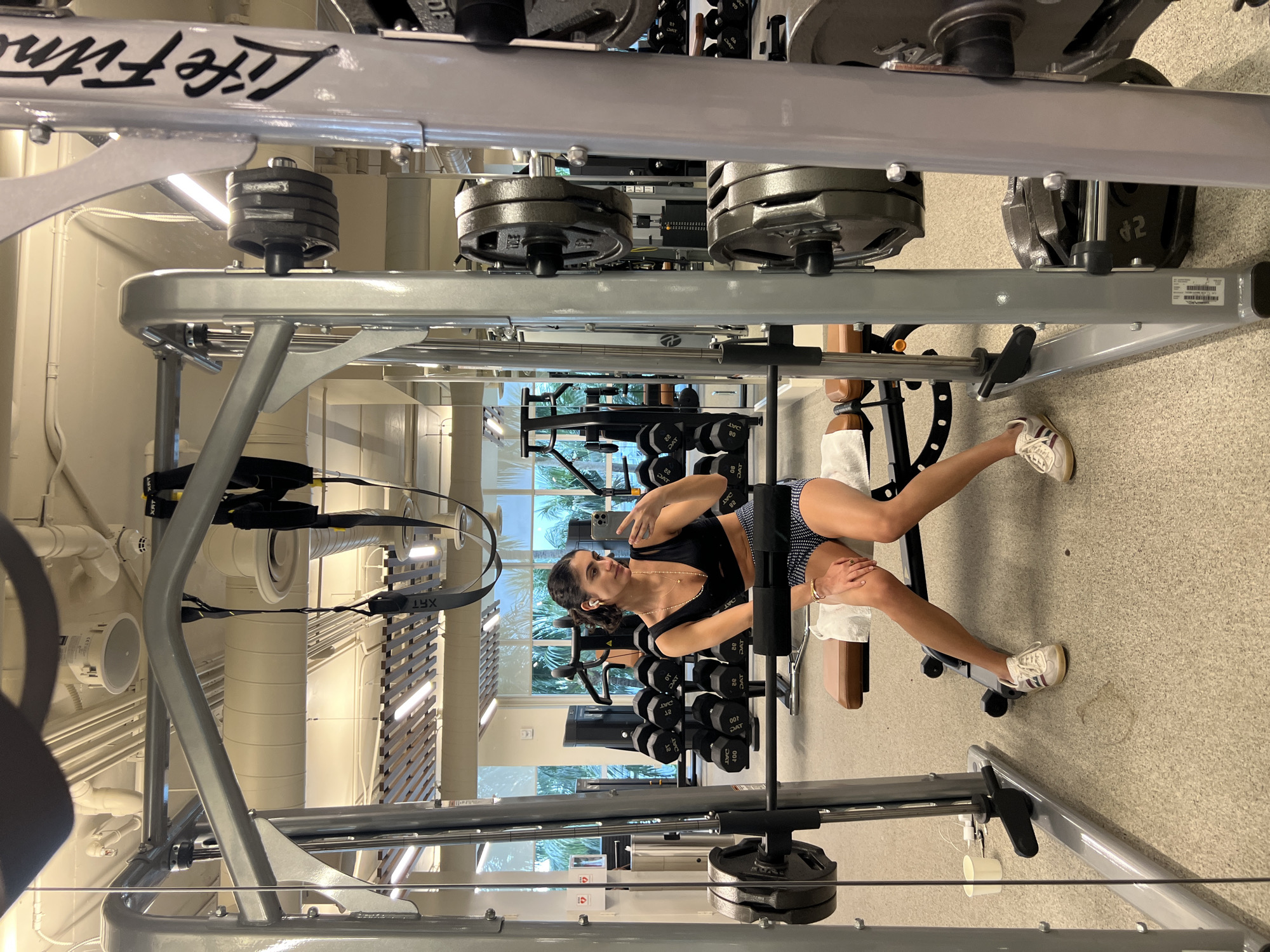 The image shows a woman taking a selfie at a gym in Miami Beach, Florida. The gym features weightlifting equipment, including a Life Fitness machine and dumbbells. The woman is dressed in athletic attire. Outside the windows, palm trees and other vegetation are visible, indicating a tropical environment. The lighting in the gym is bright, illuminating the various pieces of equipment and the woman.