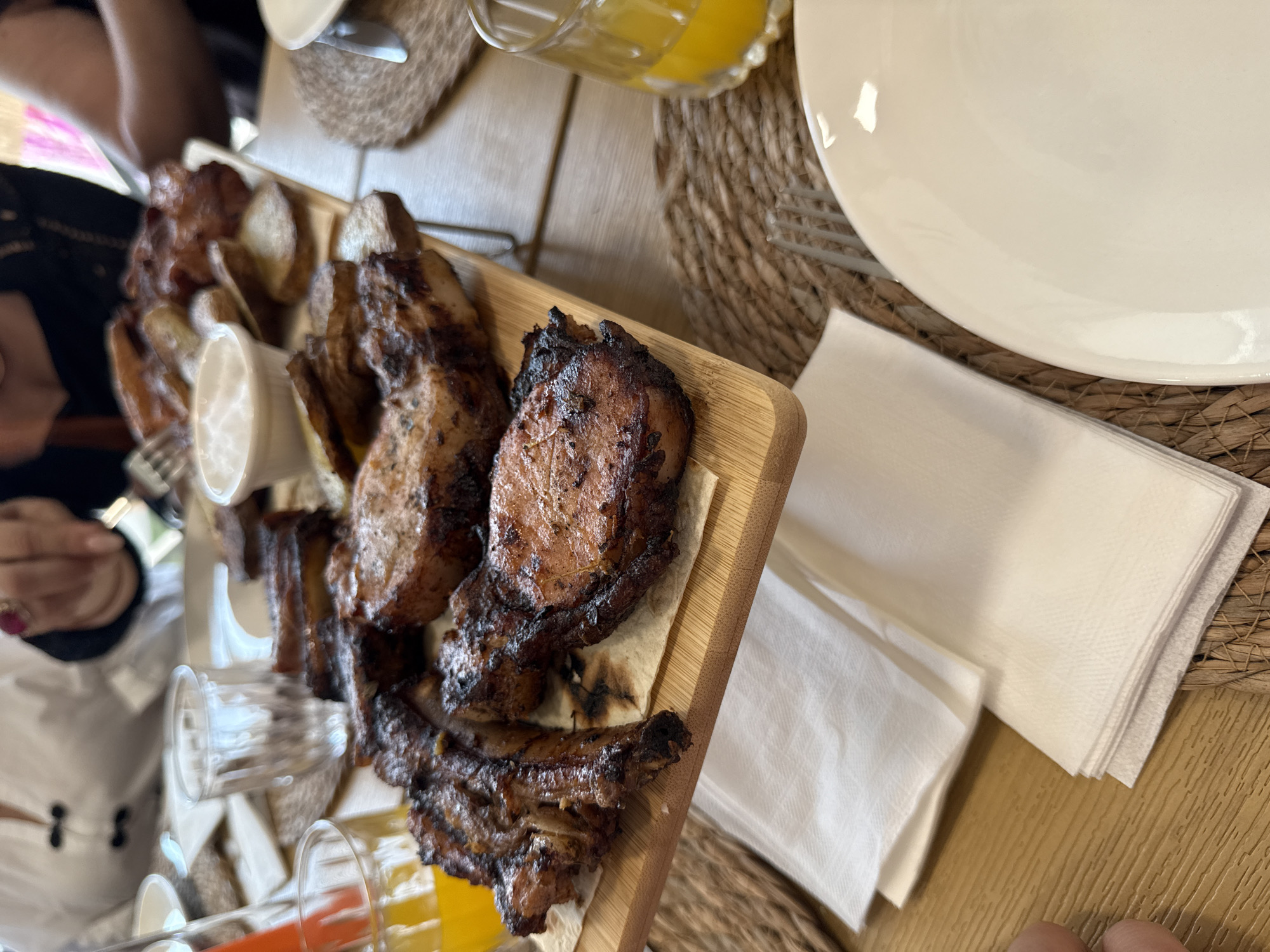 The image depicts a close-up, eye-level view of a wooden platter filled with grilled meats and roasted potato wedges. The platter is presented on a table set with placemats, white napkins, and drinks. In the background, out of focus, are individuals seated at the table. To the left, a glass contains an orange beverage, likely juice. A small white bowl holding a creamy dip or sauce is situated amongst the food on the platter. The meats appear to be pork chops and ribs, showing char marks from grilling. The overall setting suggests a meal at a restaurant, possibly outdoors given the natural lighting. The image was taken in Proshyan, Armenia.