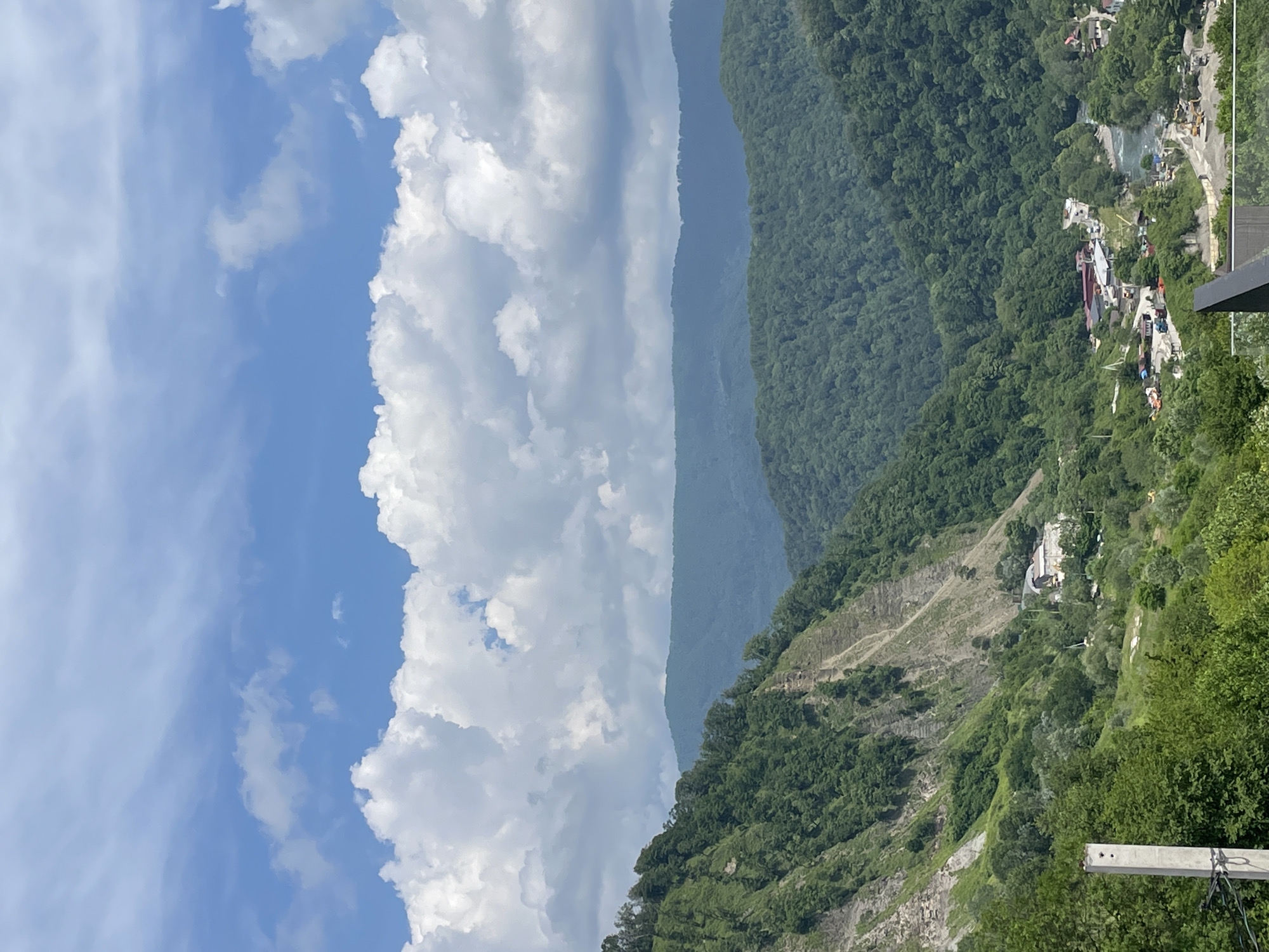 This image presents a panoramic view of a mountainous valley in the vicinity of Baranovka, Russia, under a partly cloudy sky. The upper two-thirds of the frame is occupied by a blue sky featuring numerous white cumulus clouds, ranging from small scattered formations to large, billowy masses.
The landscape below is characterized by densely forested mountains. In the mid-ground, a steep, verdant slope on the left shows exposed rock faces and areas of visible erosion or landslide scars, interspersed with dense green trees. Successive layers of mountains recede into the distance, displaying varying shades of dark green to bluish-green, indicative of atmospheric perspective and extensive tree cover.
In the valley floor, a settlement or industrial area is visible alongside a winding, light blue-green river. Numerous structures, predominantly buildings with light-colored roofs, are clustered near the river. Significant ground disturbance and what appears to be ongoing construction activity are evident, with several yellow heavy machinery (e.g., excavators, loaders) and trucks present, along with piles of aggregates. A network of roads or paths traverses this area. Some smaller residential structures with red roofs are also discernible.
In the immediate lower-left foreground, a vertical, light grey concrete utility pole with attached dark lines is partially visible. The lower-right foreground shows the edge of a dark railing and a transparent glass panel, suggesting the photograph was taken from an elevated observation point or balcony. No individuals or specific human interactions are clearly discernible within the frame.
