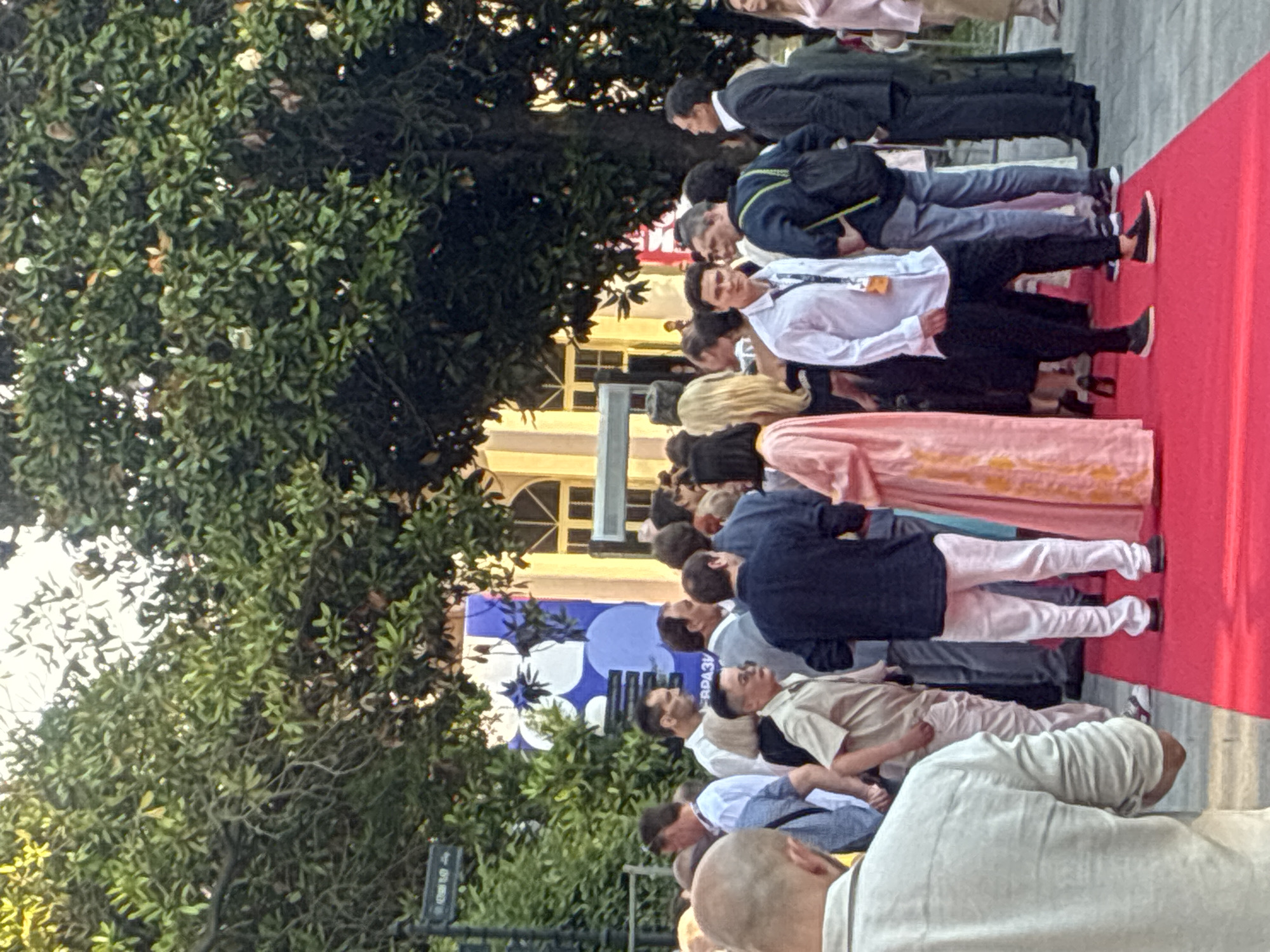 A crowd of people are standing on a red carpet in front of a building with yellow accents. The people are dressed in formal and semi-formal attire, with many men wearing suits and women in dresses. A large tree with green leaves is visible in the background, casting shadows on the scene. The setting appears to be an outdoor event, possibly a film festival or premiere, given the red carpet and the presence of numerous attendees. The location is the city of Sochi, Russia, as indicated by context not present in the image itself.