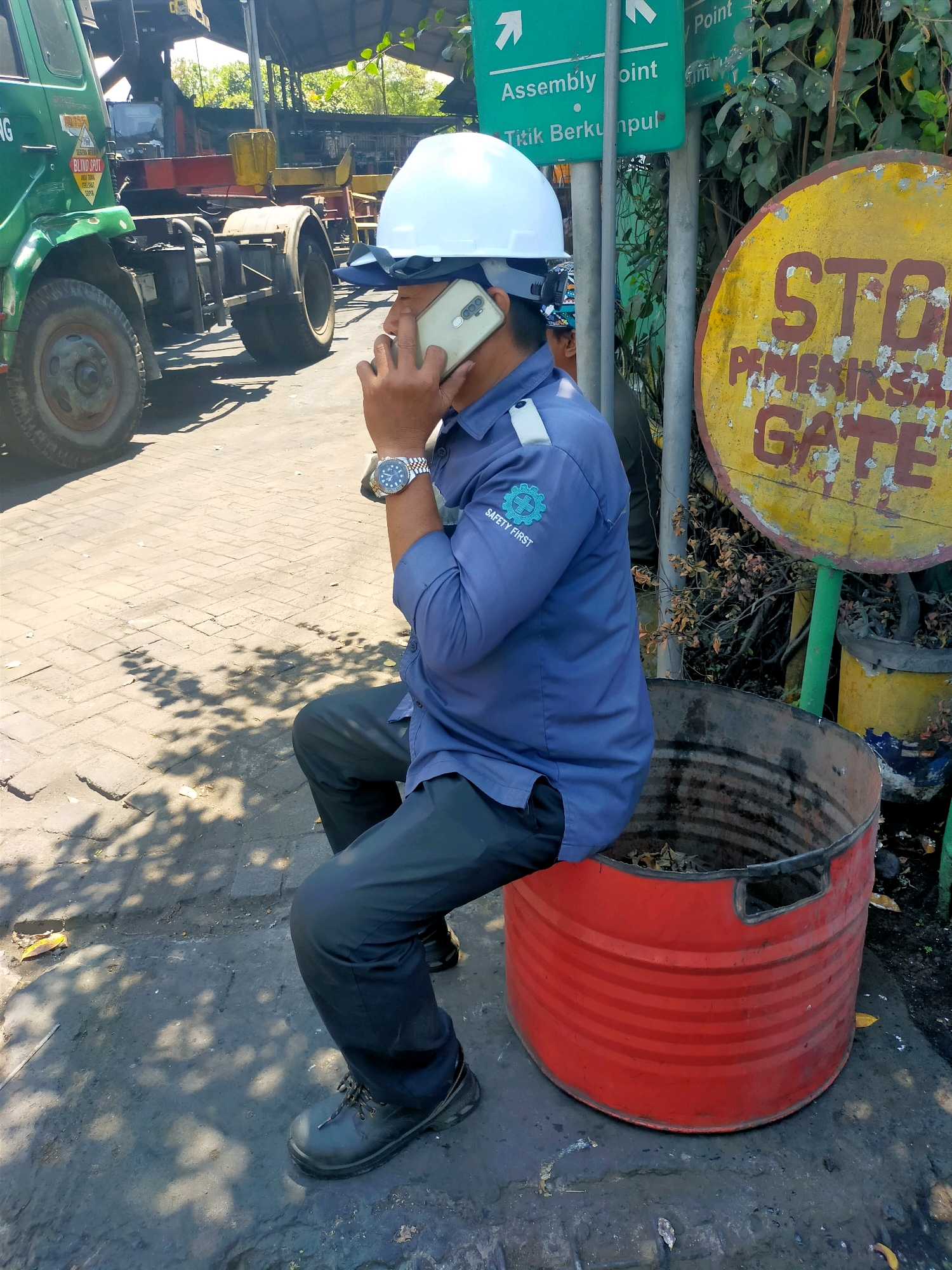 A man wearing a blue shirt, black pants, and a white hard hat sits on the edge of a large red metal drum, holding a smartphone to his ear. He wears a wristwatch on his left wrist. Behind him, to the left, is a green truck with the words "BLIND SPOT" printed on its side. To the right of the man, a circular yellow sign with red lettering reads "STOP PEMERIKSAAN GATE". The ground is paved with interlocking bricks and is covered in dust and debris. The scene appears to be outdoors, possibly in an industrial or storage area, with foliage visible in the background. The lighting suggests it is daytime, with shadows cast by unseen objects.