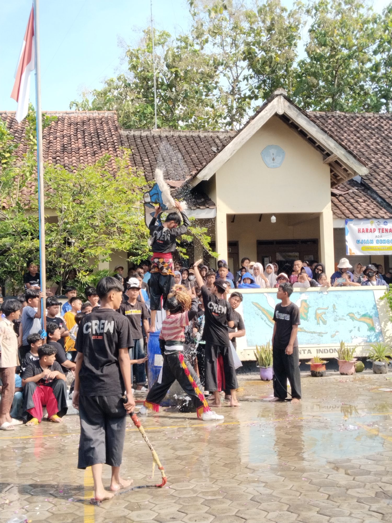 The image depicts a celebration in Karangmalang, Indonesia, on a paved area that appears to be wet. There's a group of people gathered, with many wearing black shirts labeled "CREW EST 2023." One individual is standing on top of a blue barrel, with others around him. He is pouring water over himself. A crowd of onlookers is observing from behind a low wall featuring a mural. The environment includes a building with a tiled roof, and an Indonesian flag visible in the background. There are also trees and other plants in the vicinity. The overall scene suggests a festival or community event.