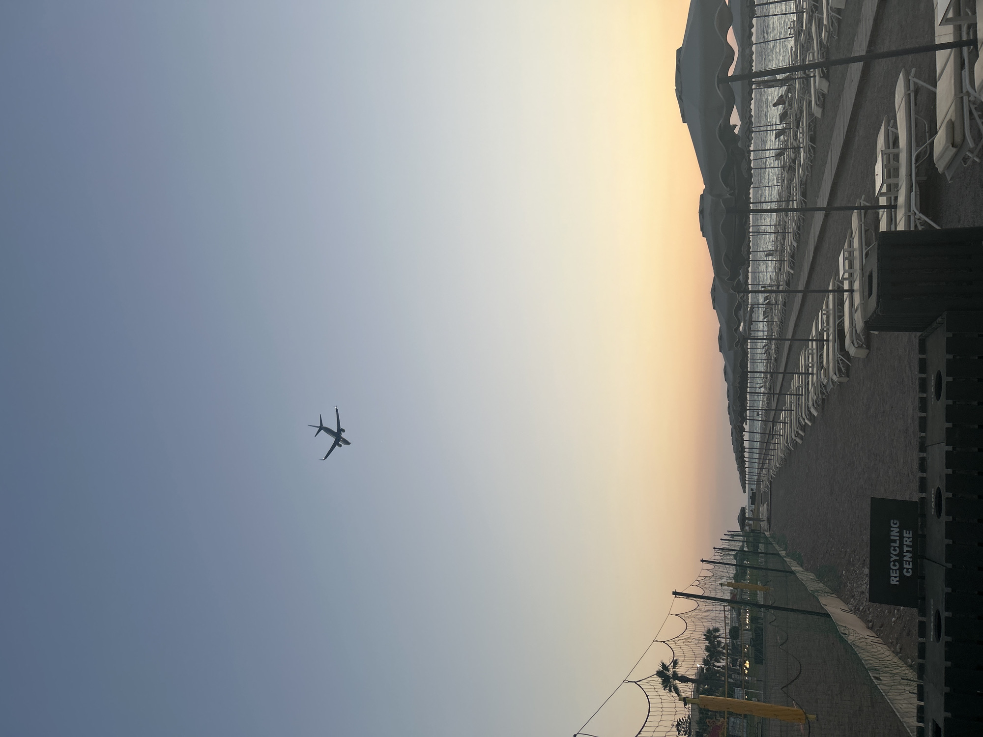 This media file captures a serene beach scene at dusk in Rhodes, Greece. The primary subjects include a row of empty beach chairs and umbrellas lining a sandy path, hinting at a day of relaxation that has concluded. In the sky, a silhouetted airplane is visible, appearing to be in its ascent or descent, adding a sense of travel and transition to the tranquil setting. The scene is bathed in the warm, soft light of sunset, with a gradient of colors from pale blue to a gentle yellow and orange on the horizon.
The overall atmosphere is peaceful and quiet, suggesting a calm evening. The location is identifiable as a beach, likely a resort area, given the organized arrangement of beach amenities. The gentle waves of the sea are visible in the background, further confirming the coastal setting. A volleyball net is also present, implying recreational activities might take place there during the day.
A notable detail is the "RECYCLING CENTRE" sign visible on a dark structure on the right side of the image. The weather appears clear and pleasant, characteristic of an evening in a Mediterranean climate. The time of day is clearly sunset. No people are visible in the frame, emphasizing the quiet and end-of-day feel. The composition creates a sense of vastness with the open sky and the expansive beach, contrasted with the linear arrangement of the beach furniture.