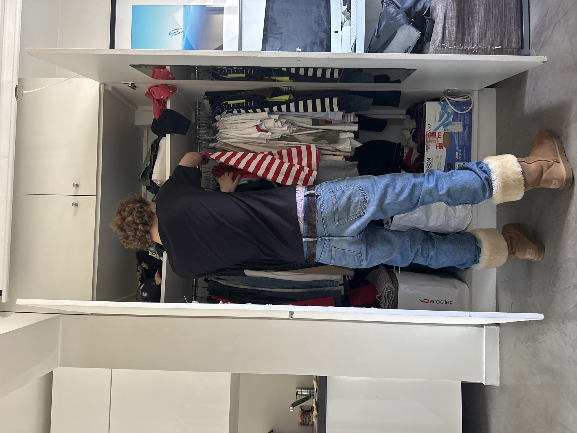 A person is reaching into a partially open closet in an interior space. The individual has curly brown hair and is wearing a black t-shirt, blue jeans with a brown belt, and tan Ugg boots with fur trim. The closet is white and full of clothing, including striped shirts and items with red and white stripes. A blue box is visible in the closet. There's a sink with a faucet visible near the bottom of the frame. An abstract print is on top of the closet. The scene is indoors in what appears to be a modern home. No information is given to associate it with Paris, France.