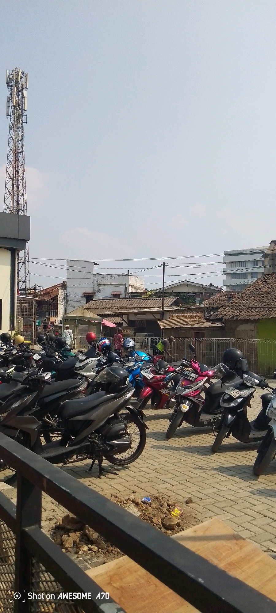 The image captures a street scene in Gadobangkong, Indonesia, on a clear day. In the foreground, a black metal railing partially obscures the view of a paved area filled with parked motorcycles. The motorcycles are varied in color and style, with some displaying helmets on their seats or handlebars. Some of the motorcycles are dark gray, black, blue, pink, and red. Behind the motorcycles, several buildings of different architectural styles are visible, including a white multi-story building on the right and a lower, older structure with a tiled roof to its left. A tall cell tower stands prominently on the left side of the image, near a building with a gray facade. The ground is paved with interlocking bricks, and there are piles of dirt and debris scattered in some areas. The sky is mostly clear with a few faint clouds. There are a few individuals visible in the background, some standing near the buildings. The overall impression is of an outdoor public space, possibly a parking lot adjacent to a commercial or residential area.