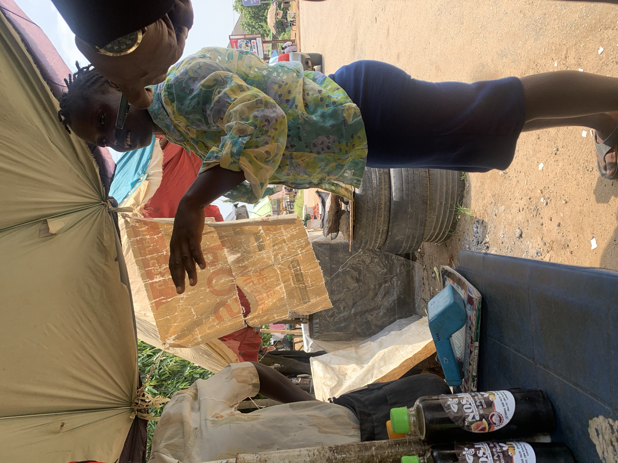 In an outdoor market stall in Abuja, Nigeria, a Black woman with braided hair and wearing a colorful floral-patterned blouse and a dark blue skirt stands under a tent. She is looking towards her right, with her mouth slightly open in a smile or speech. Her right hand is extended, and she appears to be holding a small object, possibly a pen or stylus, near her face. A portion of another person's arm, wearing a gold-colored watch, is visible on the right side of the frame, reaching towards the woman. In the background, a man in a light-colored shirt and dark pants is partially visible behind a stack of materials. On a table in the foreground, two bottles of a dark liquid with labels featuring bees are displayed, alongside a blue card reader on top of some papers. The environment is dusty, with piles of tires visible behind the stall. The scene suggests a commercial transaction or interaction is taking place.