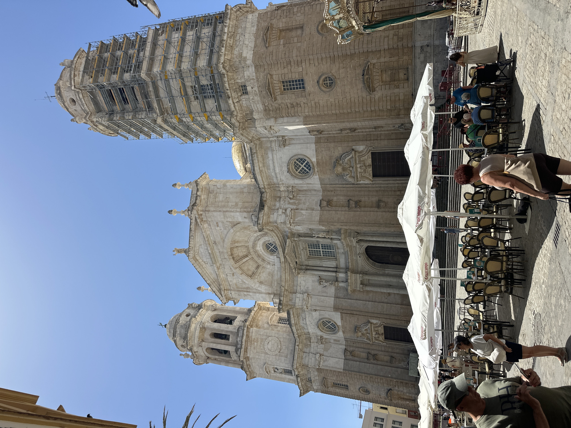 The image captures a sunlit daytime scene in Cádiz, Spain, featuring the Catedral de Cádiz, a prominent landmark. The cathedral, constructed from light-colored stone, displays architectural elements including a large dome, a bell tower with multiple arches, and ornate facades. One of the cathedral's towers is partially covered in scaffolding, indicating ongoing renovation work. In the foreground, a street cafe is set up with numerous white umbrellas providing shade over tables and chairs arranged on a cobblestone plaza. Several people are visible, some seated at the tables, others standing or walking through the area. One man in the left foreground wears a green cap and is looking at his phone. A woman in a white top and dark skirt stands near the tables, looking up. To the right of the cafe, a section of a carousel is visible, with decorative paintings on its panels. The sky is a clear, bright blue.