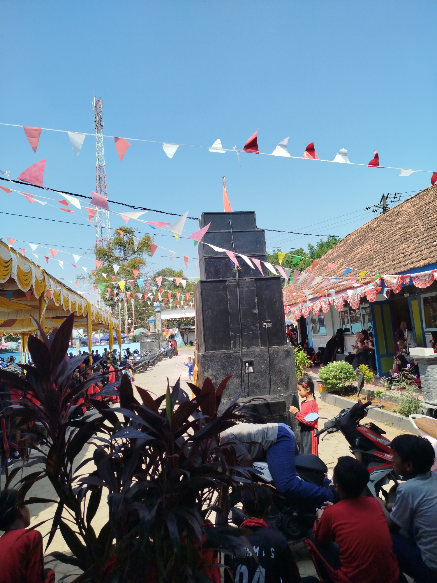 The image depicts an outdoor scene in Parseh, Indonesia, likely during a festive event, as indicated by the abundance of red and white triangular flags strung across the sky and along the building eaves. A prominent feature is a tall, multi-tiered stack of black speakers, positioned centrally in the frame. In the foreground, lush, dark red foliage provides a natural border.
The environment is characterized by a clear blue sky and a paved ground. To the left, a yellow and white tent structure is visible, suggesting a gathering area or performance space, with many people seated beneath it. A tall telecommunication tower stands in the distance, adding to the urban backdrop.
On the right side of the image, a building with a tiled roof and a blue facade is adorned with more festive decorations. Several people are gathered near the building's entrance, some seated and others standing. The street is lined with parked motorcycles.
Several individuals are engaged in various activities. A person is bent over a motorcycle, appearing to be working on it. A young girl in a red shirt stands beside the motorcycle, looking towards the speakers. A group of young boys is seated on the ground, observing the scene. Other people are scattered throughout the frame, some interacting with each other, others simply present at the event. The overall impression is one of community gathering and celebration under a bright, sunny sky.