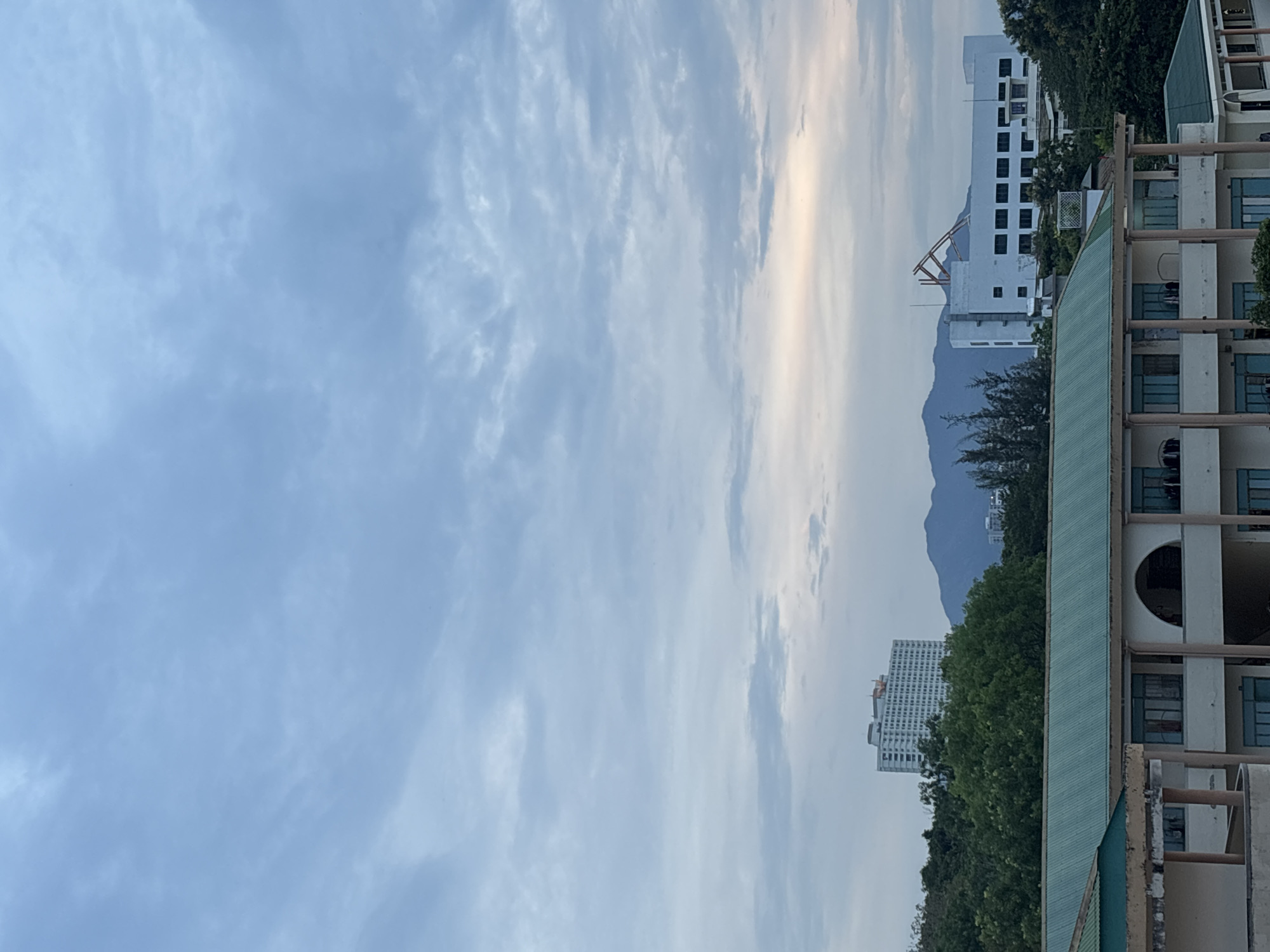 The image shows a city skyline with a cloudy sky overhead. In the foreground, there is a building with a green roof and arched windows. In the middle ground, there are trees, a tall white building, and mountains in the distance. There is also another white building with a crane-like structure on its roof. The lighting suggests it is either early morning or late evening. The scene appears to be a general view of an urban area.
