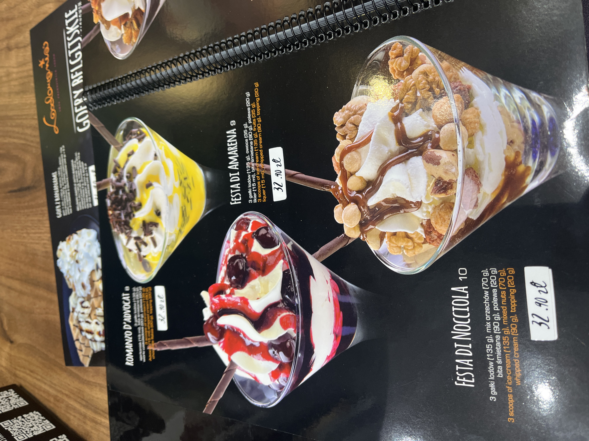 The image displays a close-up of a menu page from a restaurant. The menu features photographs and descriptions of desserts.
In the foreground, a dessert named "Festa Di Nocciola" is prominently featured. It is served in a tall, clear glass and consists of layers of ice cream, whipped cream, and a topping of mixed nuts and caramel sauce. A price tag of "32.90 zł" is visible below the description. The description lists ingredients in Polish and English, including "3 scoops of ice-cream (135 g), mixed nuts (70 g), whipped cream (90 g), topping (20 g)."
To the left, another dessert, "Romanzo D'Avocat," is partially visible. It is also served in a clear glass, layered with cream and topped with what appears to be a cherry sauce and chocolate shavings, with chocolate sticks inserted. A price tag of "32.90 zł" is also visible for this item.
In the background, a dessert named "Festa Di Amarena" is shown. This dessert is in a yellow dessert cup and contains what appears to be yellow ice cream with chocolate shavings and whipped cream. The description lists ingredients like "3 scoops of ice-cream (135 g), fruits (35 g), liquer (15 ml), whipped cream (90 g), topping (20 g)." A price tag of "32.90 zł" is visible below this description.
Further in the background, a waffle dessert with whipped cream and chocolate drizzle is partially visible at the top.
The menu itself appears to be bound with a black spiral spine. The overall background is dark, with some glare from lighting. The image is taken from a slightly angled top-down perspective. There are no people visible in the image.