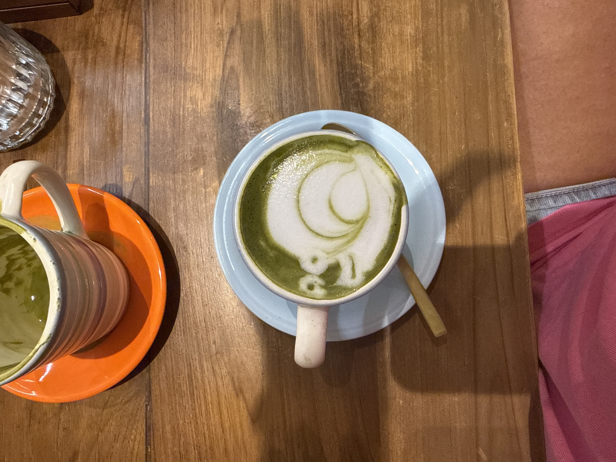The image shows a top-down view of a wooden table with a cup of matcha latte on a blue saucer, a striped mug on an orange saucer, a glass object, and part of a person wearing a pink top. The matcha latte features latte art. A wooden stirrer rests on the saucer. The image was taken in Ubud District, Indonesia.