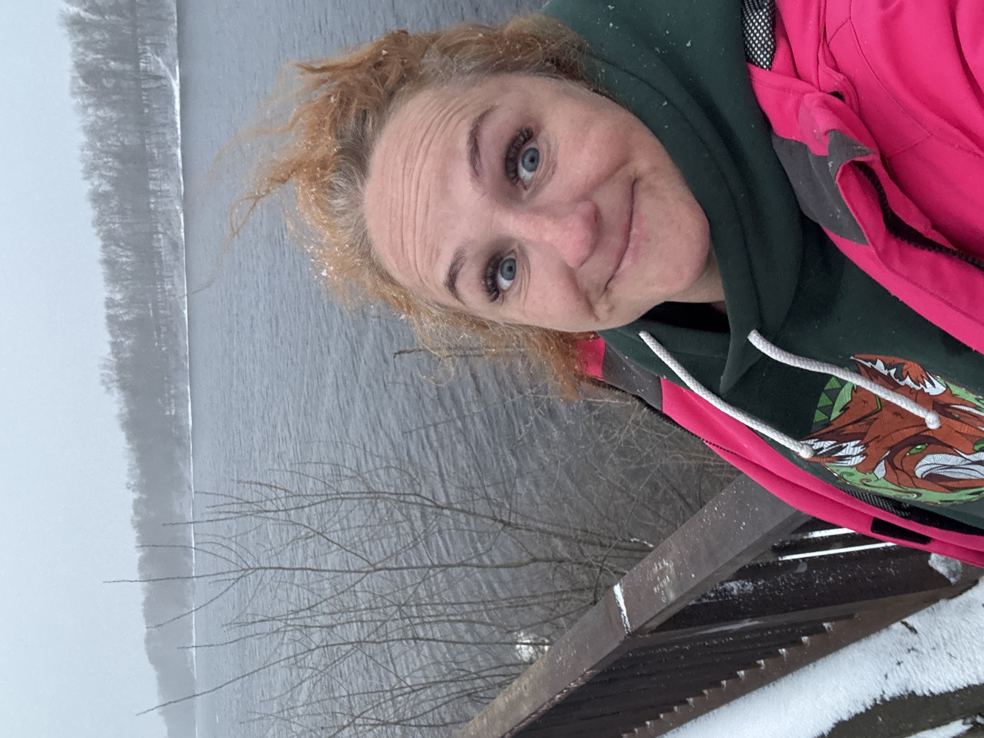 A woman with light reddish hair and blue eyes is taking a selfie outdoors. She is wearing a dark green hoodie and a bright pink jacket. Snowflakes are visible on her hair and clothing, suggesting it is snowing. The background shows a body of water (likely a river or lake) and a shoreline with trees. There is a railing covered with snow in the foreground. The location is Chaikovsky, Russia.