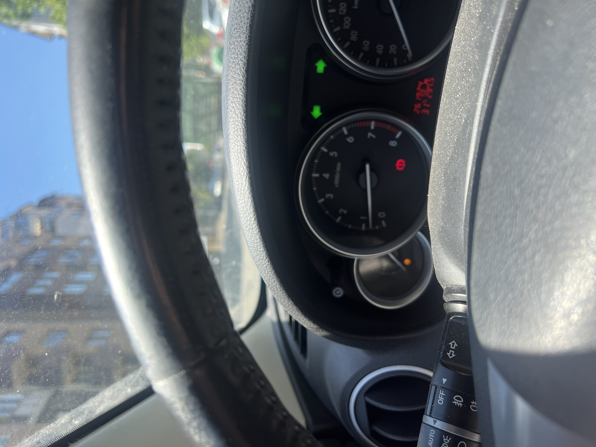 The image shows the dashboard and steering wheel of a vehicle, likely a car. The speedometer indicates the speed, while the tachometer shows the engine RPM. Several warning lights are illuminated on the dashboard, and the turn signal lever is visible. The odometer reading is "261924" and a separate number "312915". Outside the car window, a building is visible under a blue sky. It is not possible to determine if this image was taken in Burgas, Bulgaria.