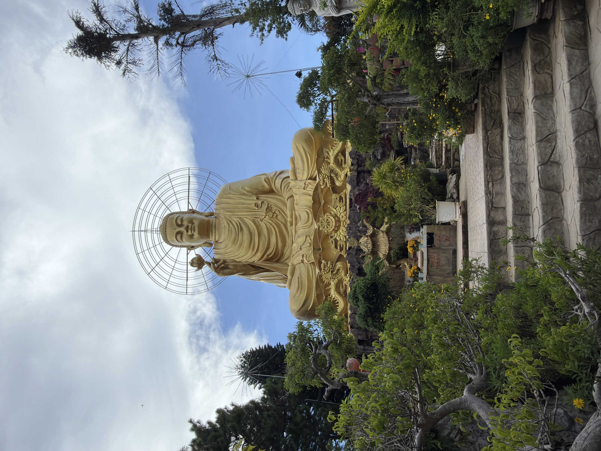 The image shows a large, golden statue of a seated Buddha holding a lotus flower. The Buddha statue is centrally located and appears to be made of metal. It is surrounded by lush greenery, including trees, bushes, and potted plants. There are stone steps leading up to the statue, and a stone pathway in the foreground. In the background, there are more trees and a cloudy sky. The statue is adorned with a circular halo behind its head. The overall scene is serene and appears to be located in a temple or a place of worship, likely in an outdoor setting within the city of Lâm Viên - Đà Lạt, Vietnam, given the mention of the location.