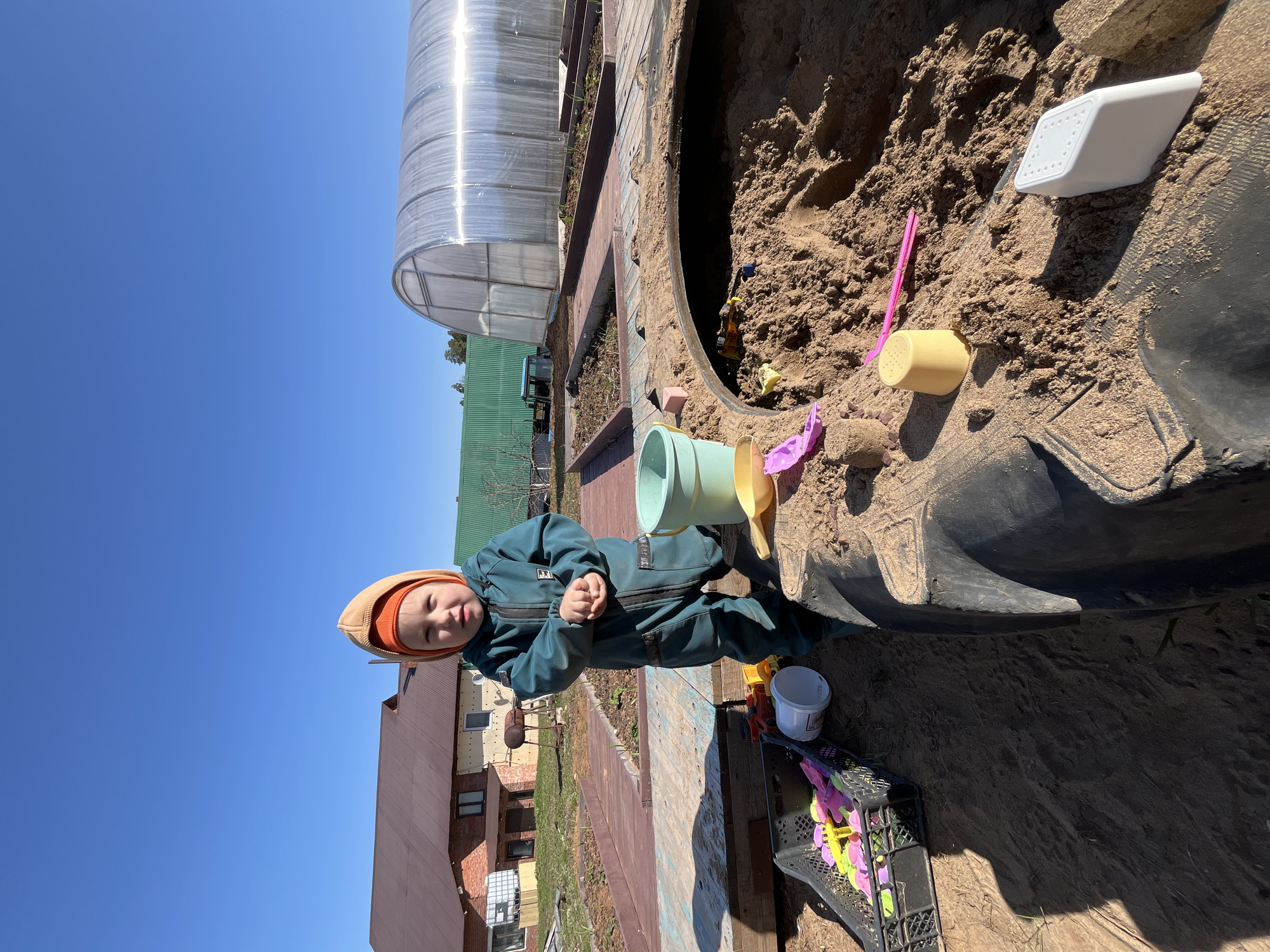 A young child in a blue jumpsuit and orange hood stands next to a large sandbox made from a black tire in Chaikovsky, Russia. The sandbox is filled with sand and various toys, including buckets, a watering can, and shovels. A greenhouse is visible in the background, along with a house under construction. The sky is clear and blue.