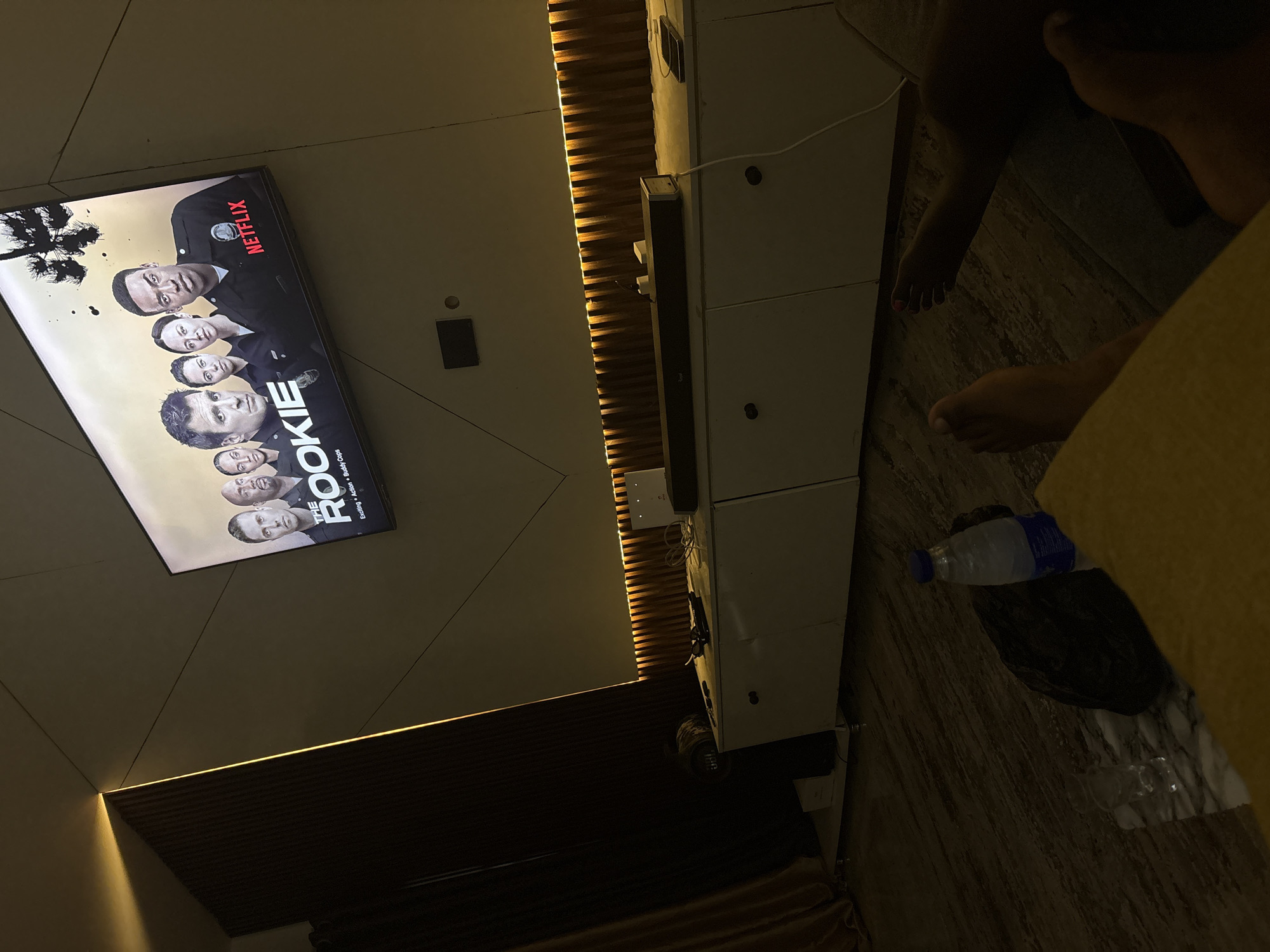 The image shows a television displaying the Netflix series "The Rookie." A partial view of a person is visible, including their feet and legs. A water bottle and a black bag are on the floor. The room appears to have modern furniture and decor, with wood paneling and a marble floor. The location, Iddo Tudunwada, Nigeria, cannot be verified from the image.