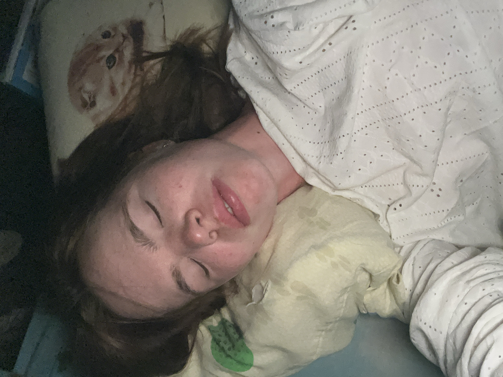 A fair-skinned person with closed eyes is lying down. They are under a white blanket with a pattern of small holes. Their head rests on a pillow that has a yellowish hue and a green circular design visible on it. There is another pillow behind their head with an image of a cat on it. The lighting in the room is dim. There is no information about location within Chaikovsky, Russia, or any details suggesting it is in Chaikovsky at all.