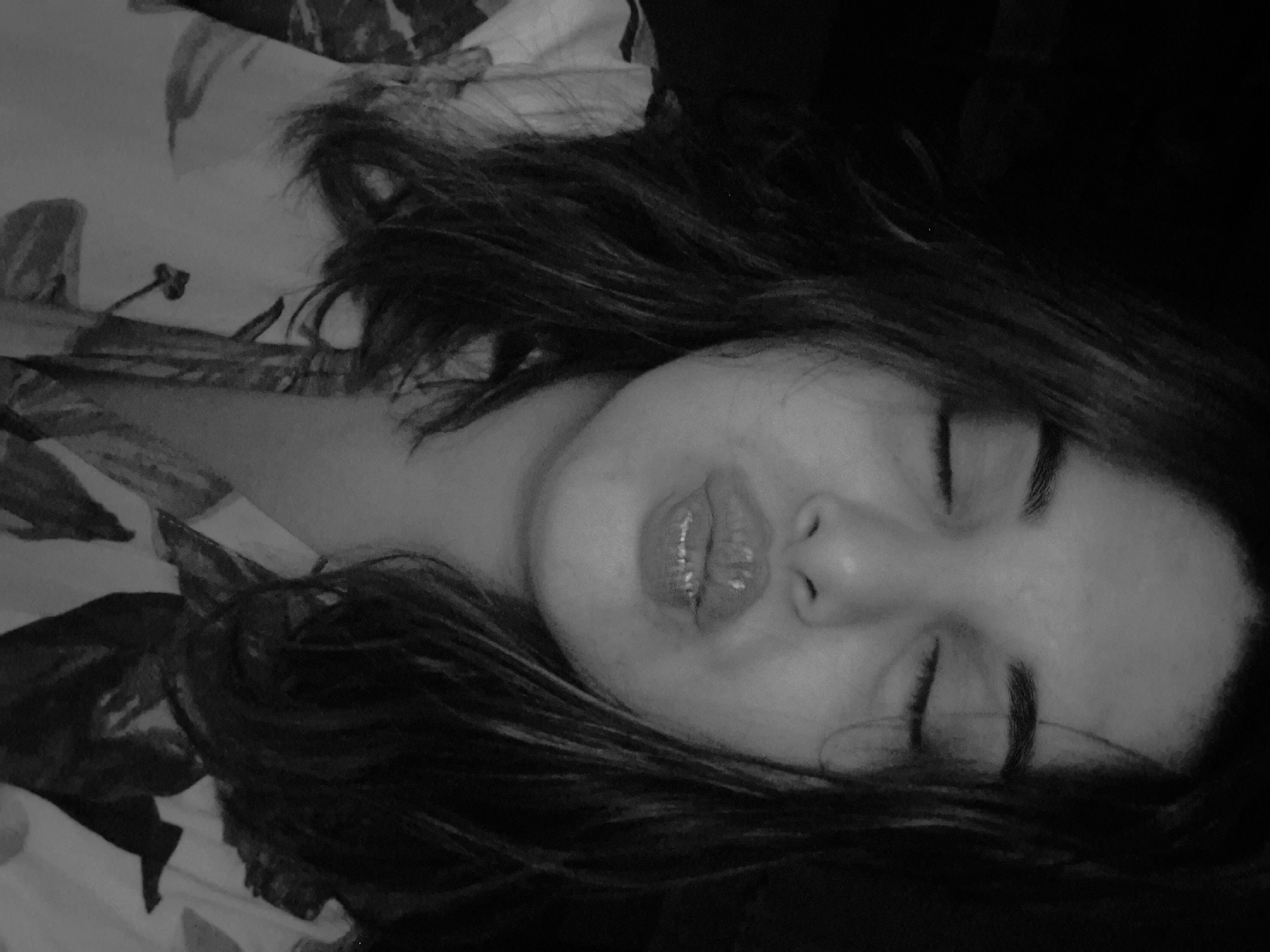 The image is a black and white photograph of a person lying down with closed eyes. The person has dark hair that surrounds their face. The person's lips are slightly parted. The person is lying on a surface with a floral pattern. There is no indication of the location being in the city of Амзя, Russia.